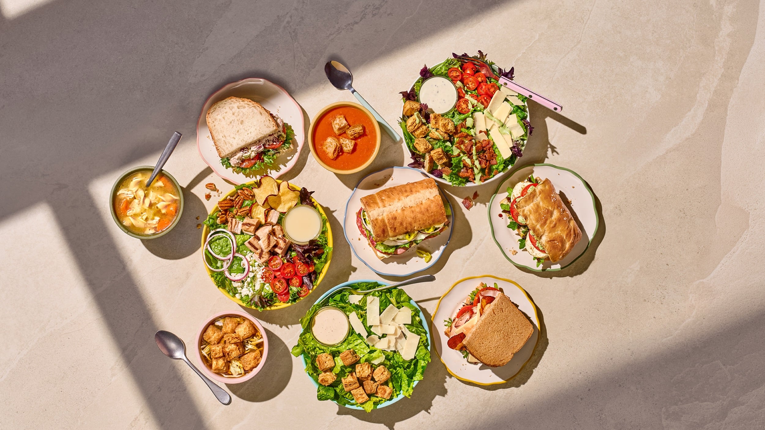 A top-down view of several bowls of salad, soup, and various sandwiches arranged on a light stone surface.