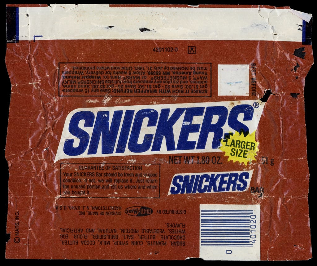 A vintage, wrinkled Snickers candy bar wrapper in brown and white, featuring the brand logo and product information.