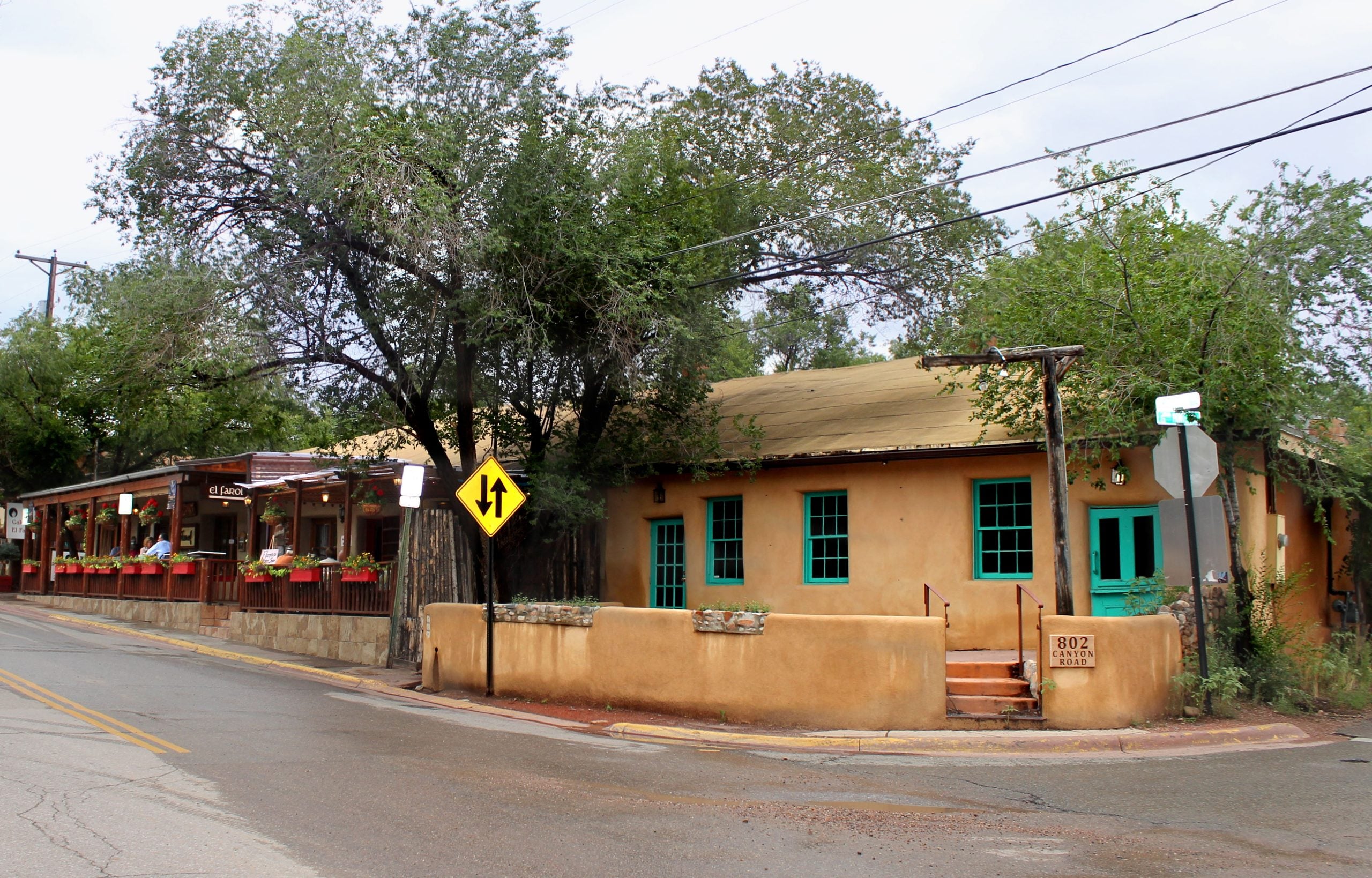 An adobe building with turquoise trim at 802 Canyon Road, next to a restaurant with outdoor seating.