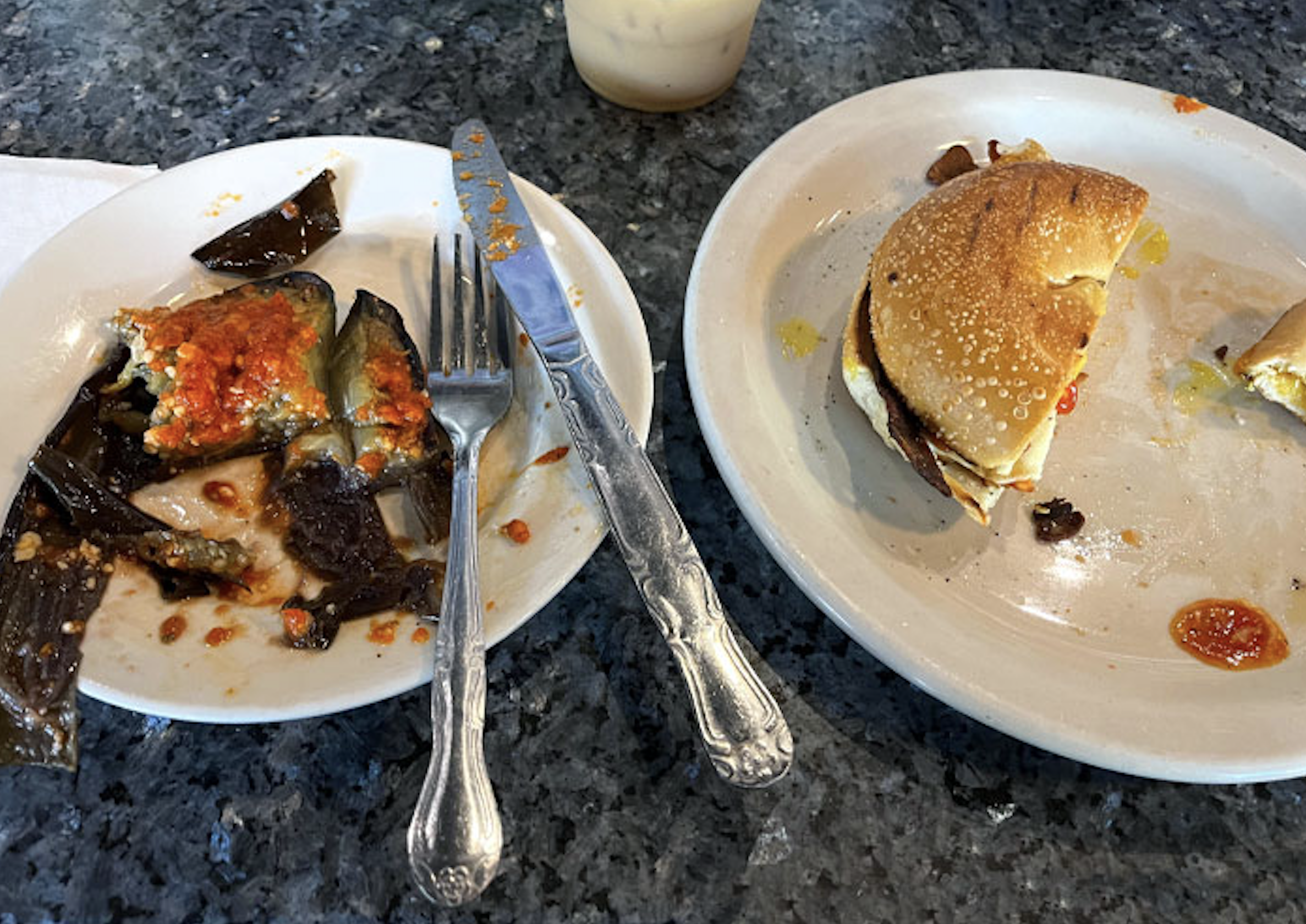 Two plates on a speckled countertop: one holds half a bagel sandwich with eggplant, the other has sliced eggplant with red sauce. A knife, fork, napkin, and drink are nearby.