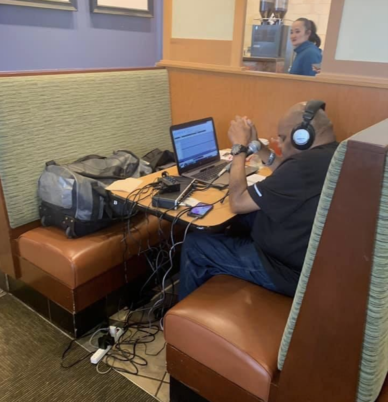 A person with headphones sits in a booth using a laptop and microphone, surrounded by cables and equipment. A woman stands in the background, smiling near a drink machine. This is one of the rare restaurant customers.