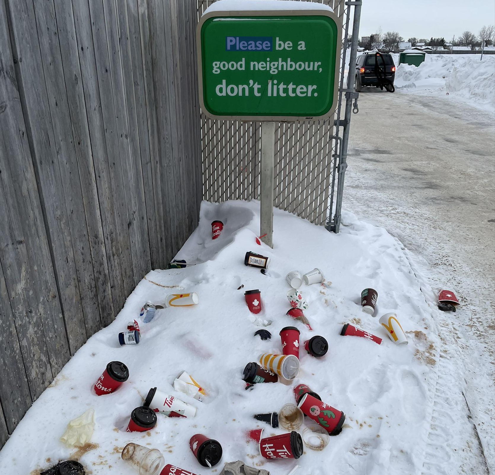 A snowy ground is littered with coffee cups and trash under a sign that reads, “Please be a good neighbour, don’t litter.” A car is parked in the background.