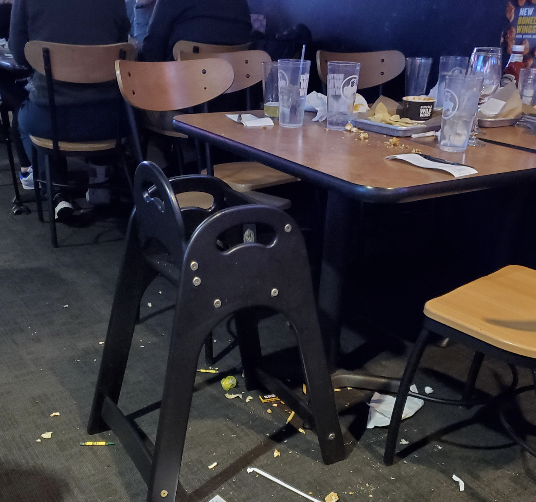 A messy restaurant scene with empty glasses, plates, and napkins on the table, food and trash on the floor, and a black high chair in the foreground.