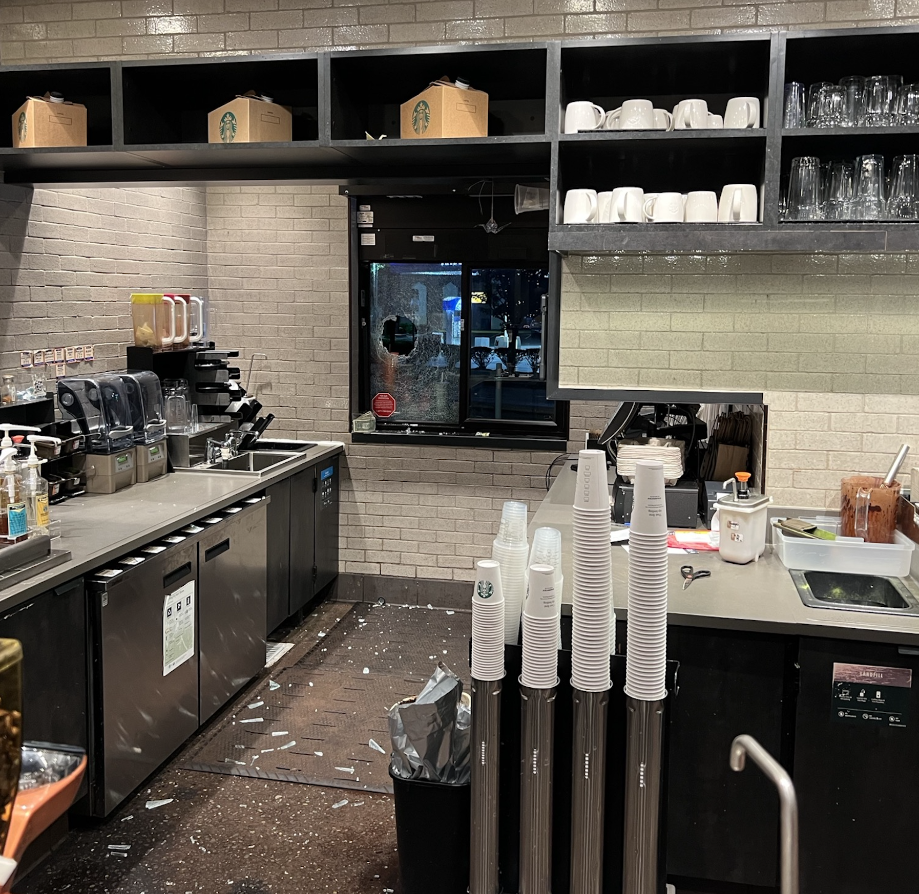 A Starbucks counter with a broken drive-thru window, shattered glass on the floor, and cups stacked near the sink; the area appears recently damaged.