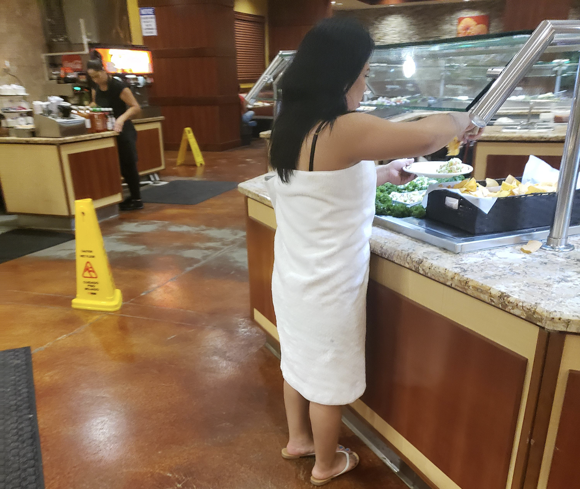 A woman wearing a white towel and flip-flops serves herself food at a buffet. Another person is in the background near a “Wet Floor” sign. The setting appears to be a casual restaurant.
