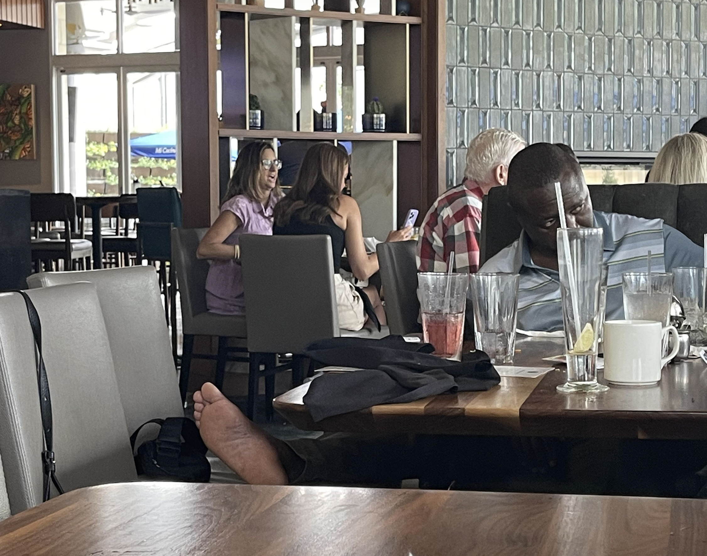 Several people sit at tables in a restaurant. Two women in the background smile and talk. In the foreground, a man in a striped shirt appears to be sleeping with drinks and empty glasses on the table.