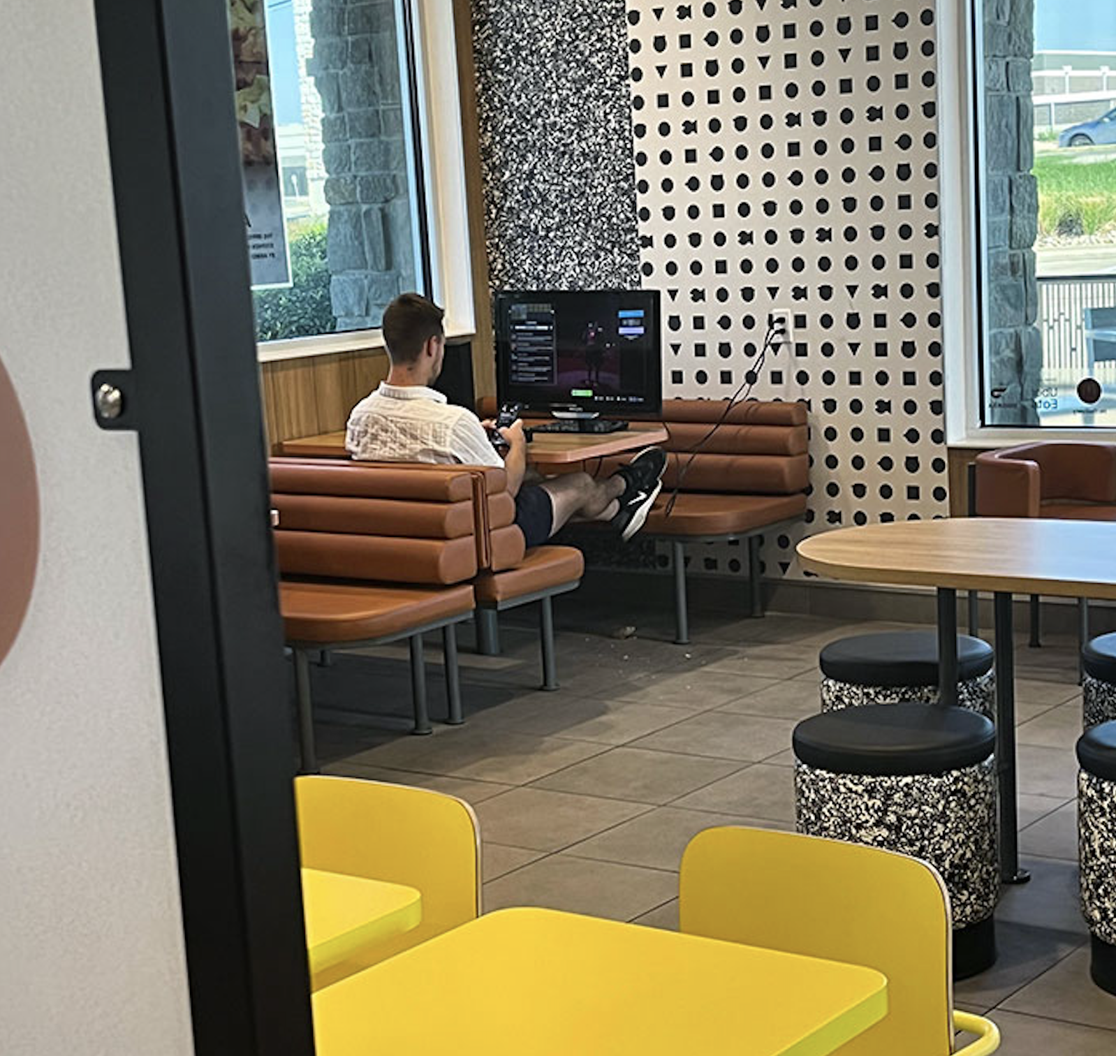 A person sits alone on a brown bench in a modern fast-food restaurant, playing a video game on a TV set up in the corner. The restaurant has yellow chairs and patterned walls.