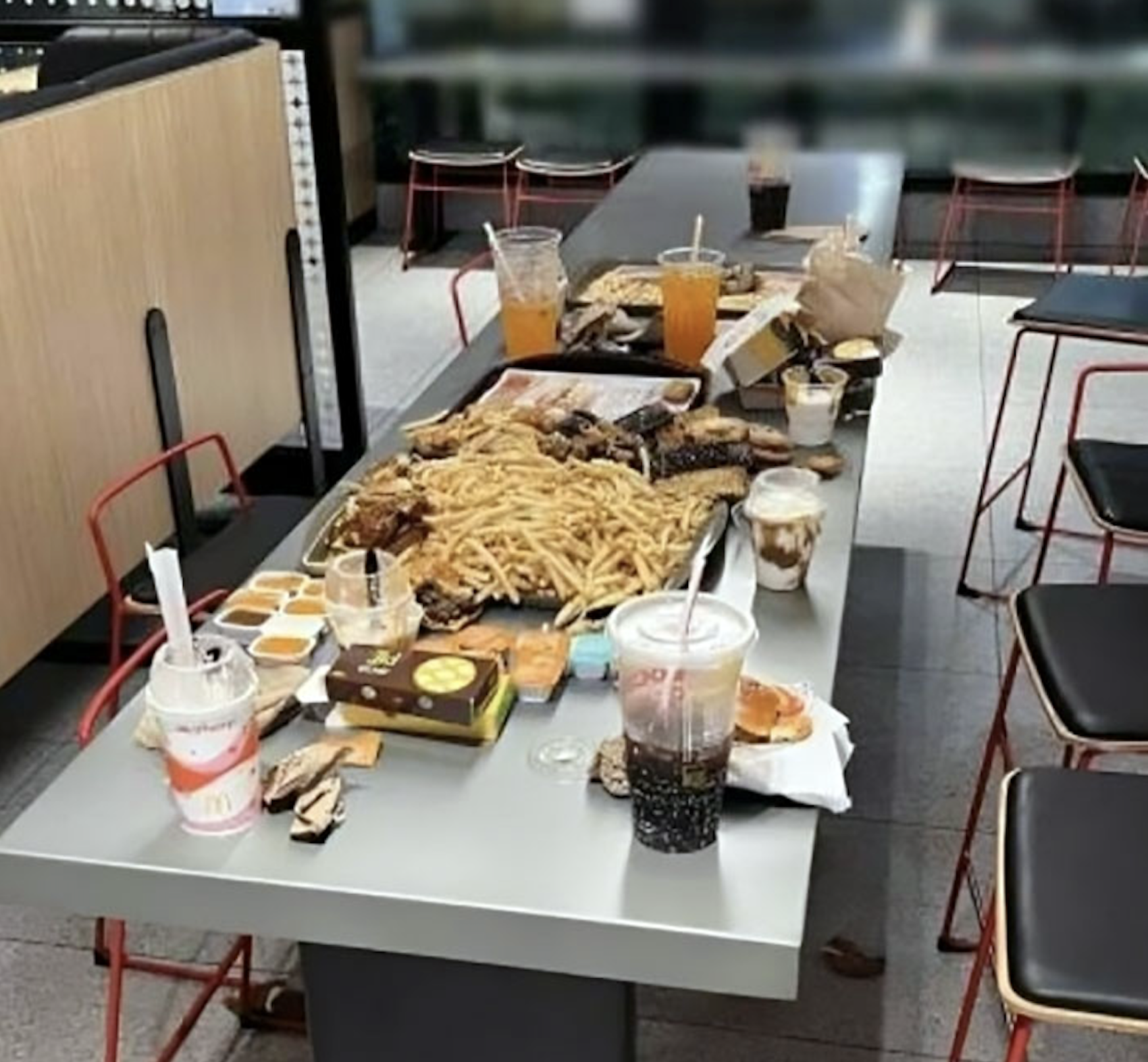 A long table in a fast-food restaurant is covered with large amounts of fries, burgers, chicken, sauces, drinks, and milkshakes, left behind by rude customers. Empty chairs surround the messy table.