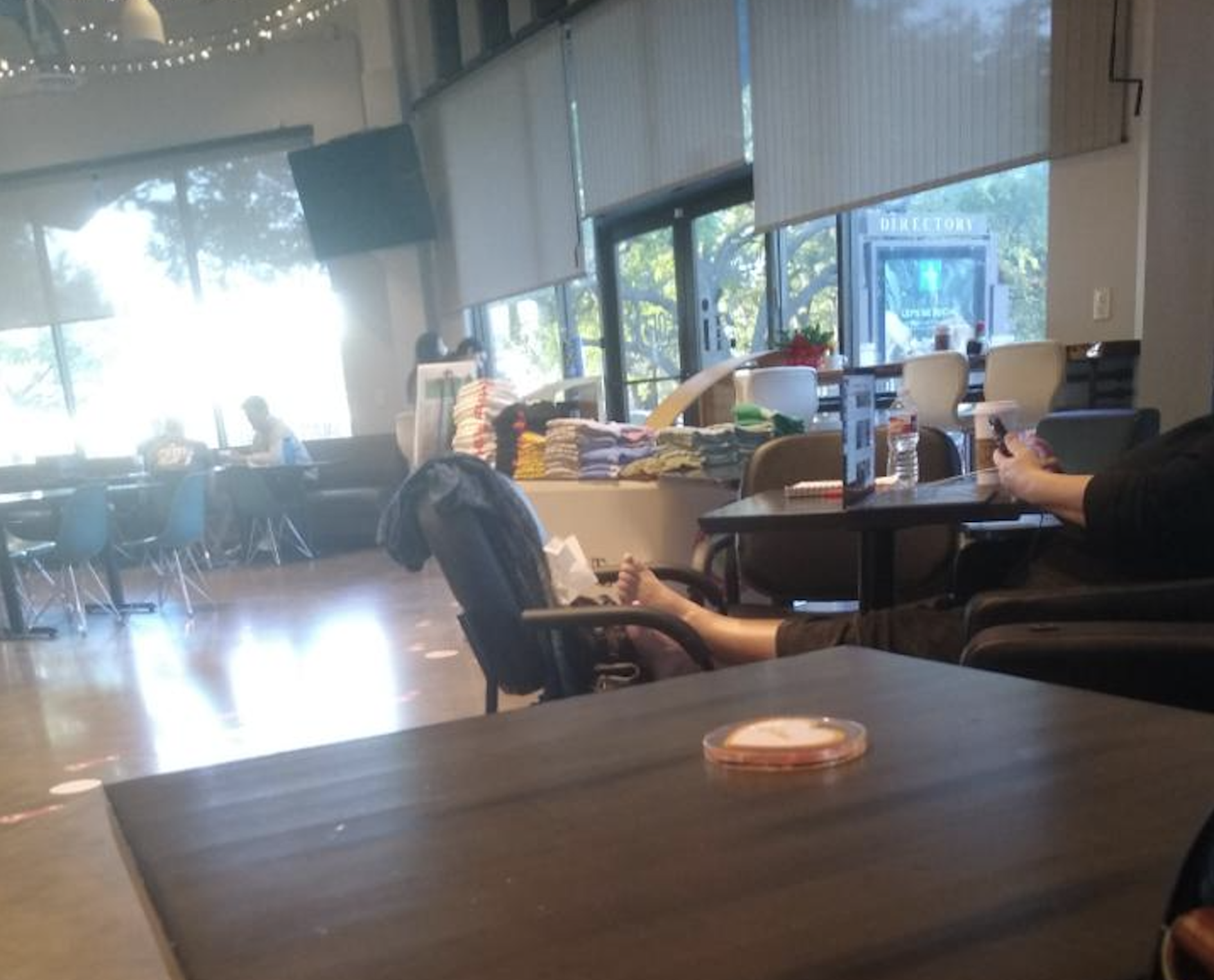 A relaxed cafe scene with people sitting at tables, one person with feet up on a chair, folded towels on a counter, large windows, and trees visible outside.