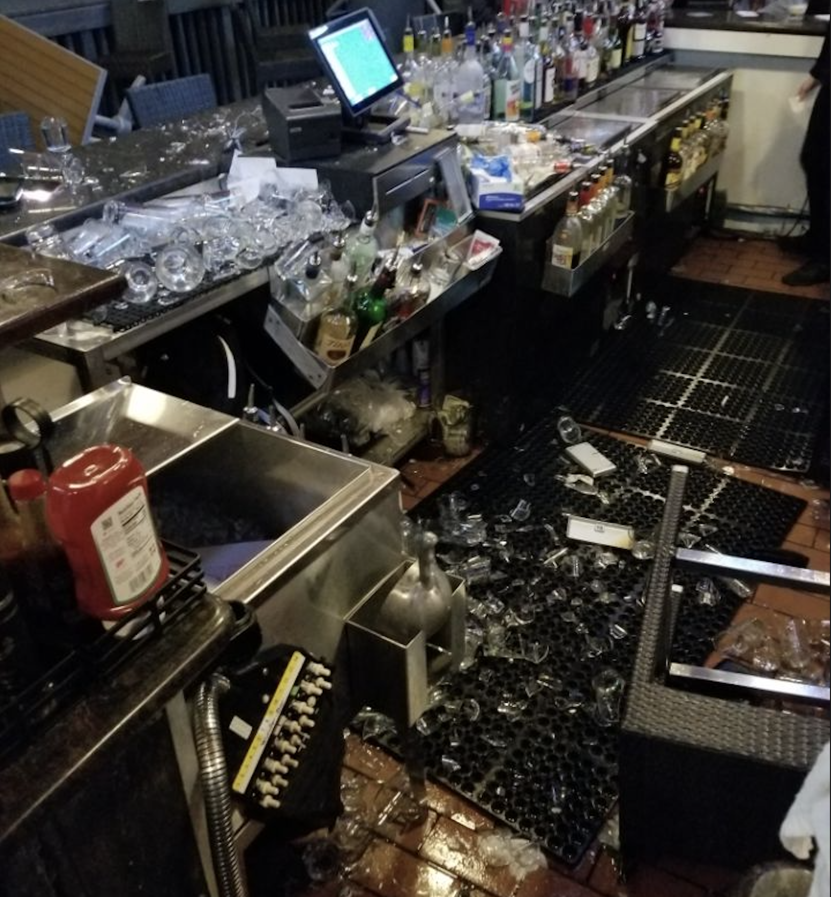 Broken glassware and bottles scattered across a messy bar floor, with many glasses fallen into sinks and onto mats. Various liquor bottles and condiments remain organized on the counter.