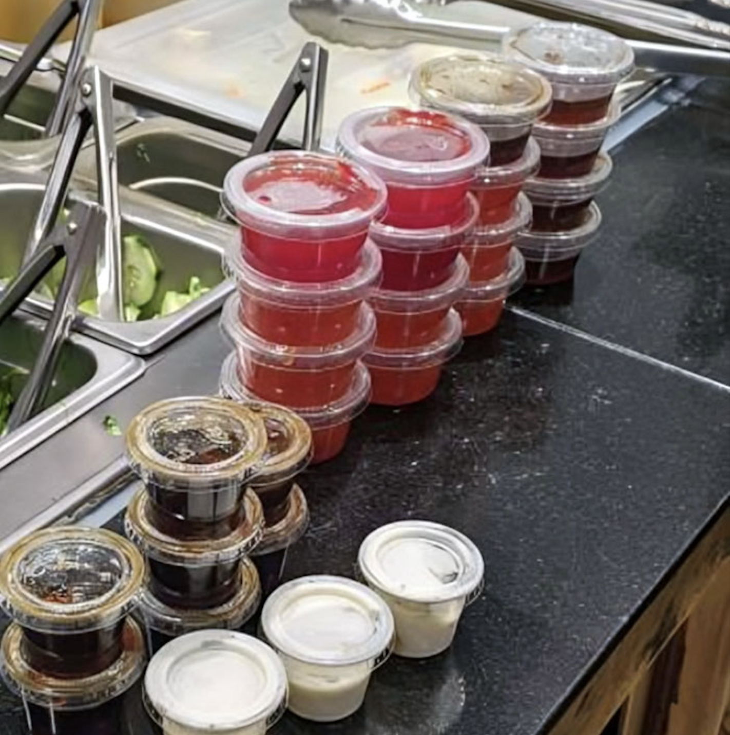 A row of small plastic sauce containers, filled with red, brown, and white sauces, stacked on a black counter beside a salad bar with metal tongs and container bins.