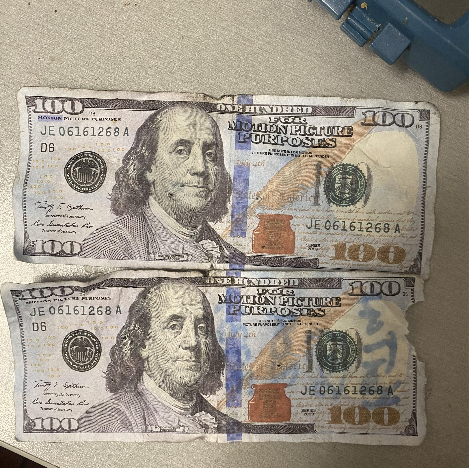 Two worn hundred-dollar bills labeled “For Motion Picture Purposes” lie on a surface. They feature Benjamin Franklin and state “This note is for motion picture purposes. It is not legal tender.” Both share the same serial number, JE 06161268 A.