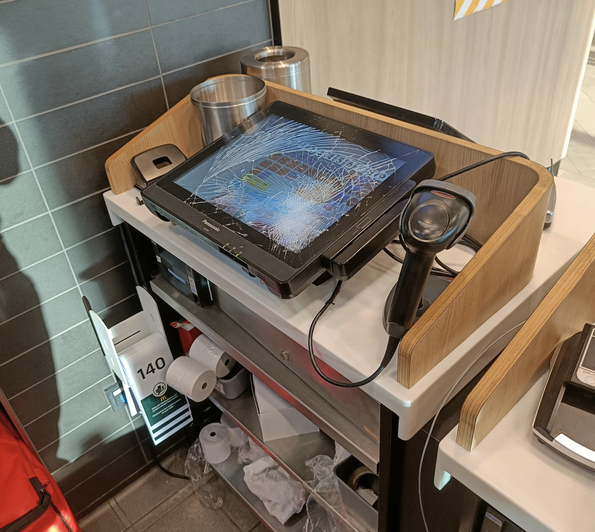 A shattered touchscreen monitor sits on a counter, surrounded by a barcode scanner and various supplies in a modern interior setting.