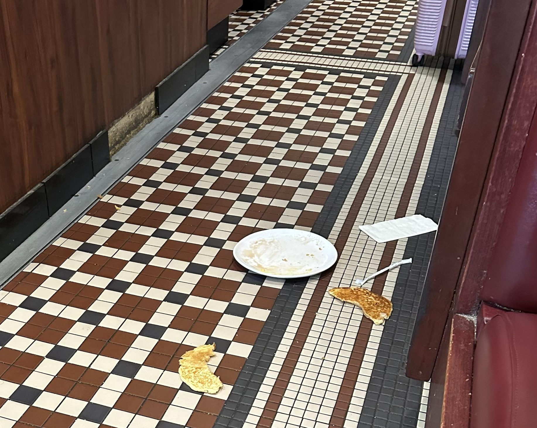 A white plate, a napkin, and pieces of food are spilled on a checkered tile floor between a booth and a wooden wall in a restaurant.