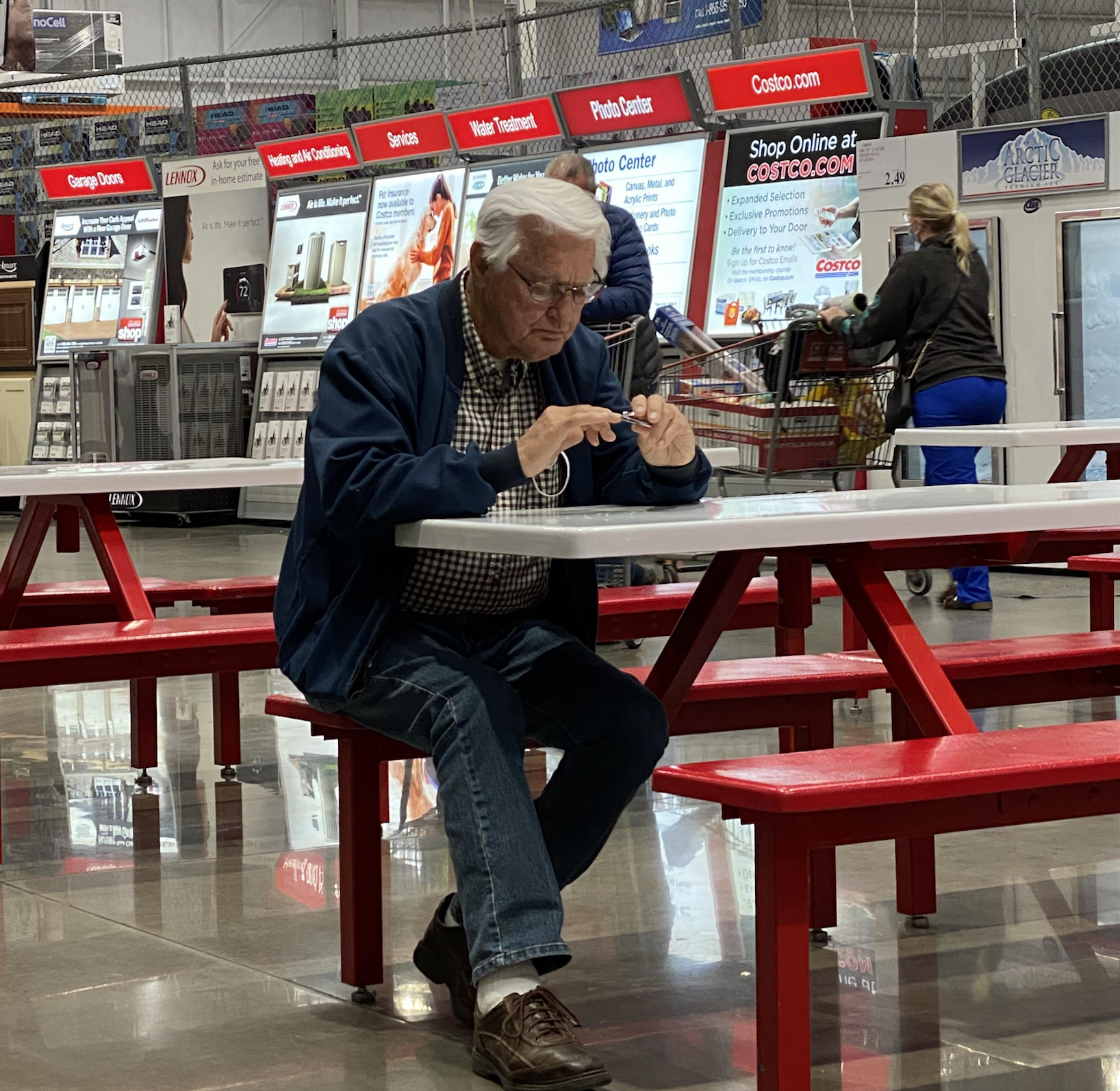 An older man sits alone at a red picnic table in a warehouse store, focused on his smartphone. Store displays and people walking by are visible in the background.