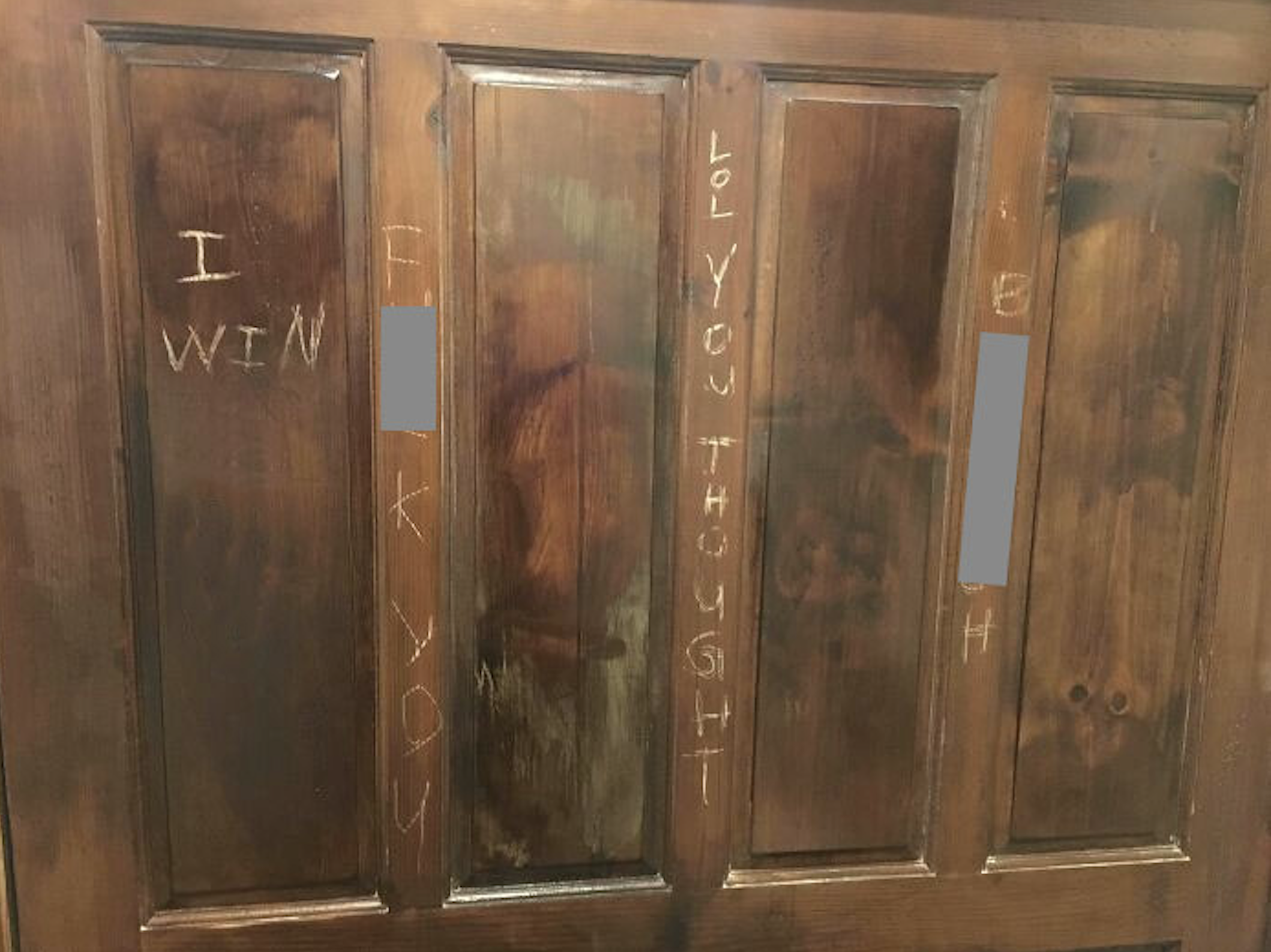 A wooden door with panels shows crude carvings, including phrases like “I WIN,” “LOL YOU THOUGHT,” and obscured profanity scratched into the surface.