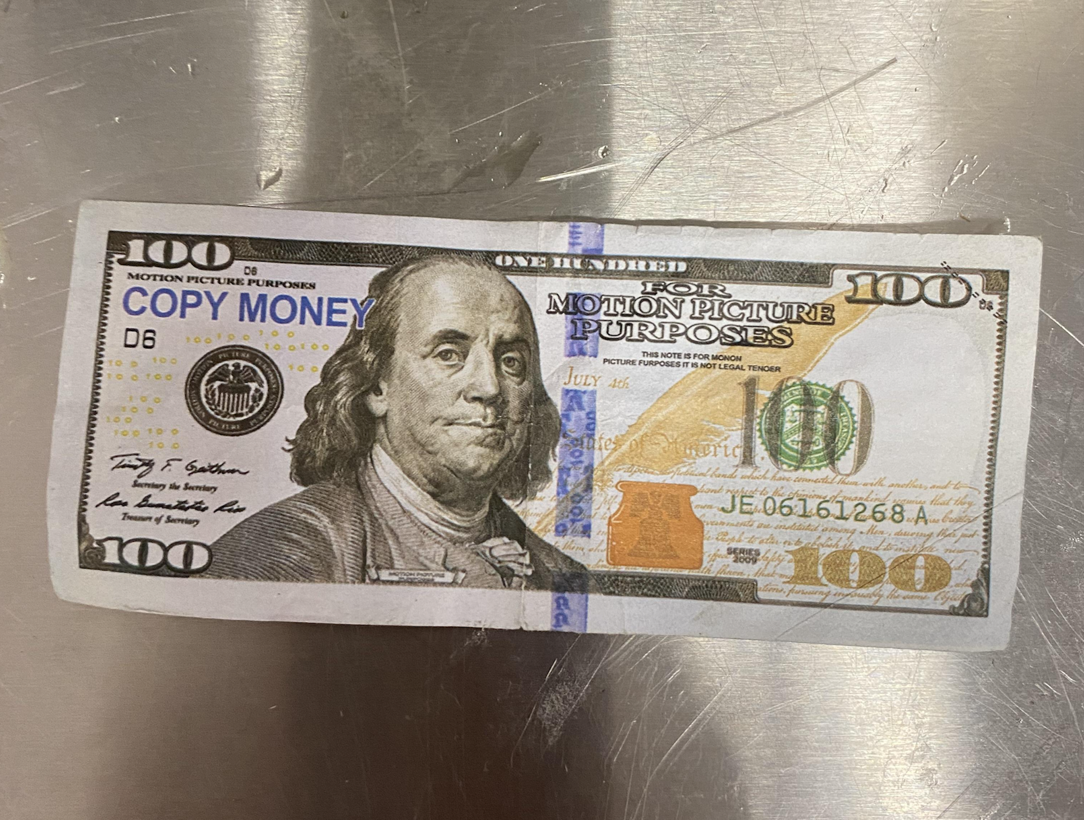 A fake $100 bill marked “COPY MONEY” and “FOR MOTION PICTURE PURPOSES” lies on a metallic surface. The bill mimics a real note but has clear labels indicating it is not legal tender.