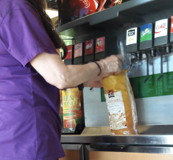 A person in a purple shirt fills a large bag of caramel popcorn from a soda fountain, with various drink dispensers visible in the background.