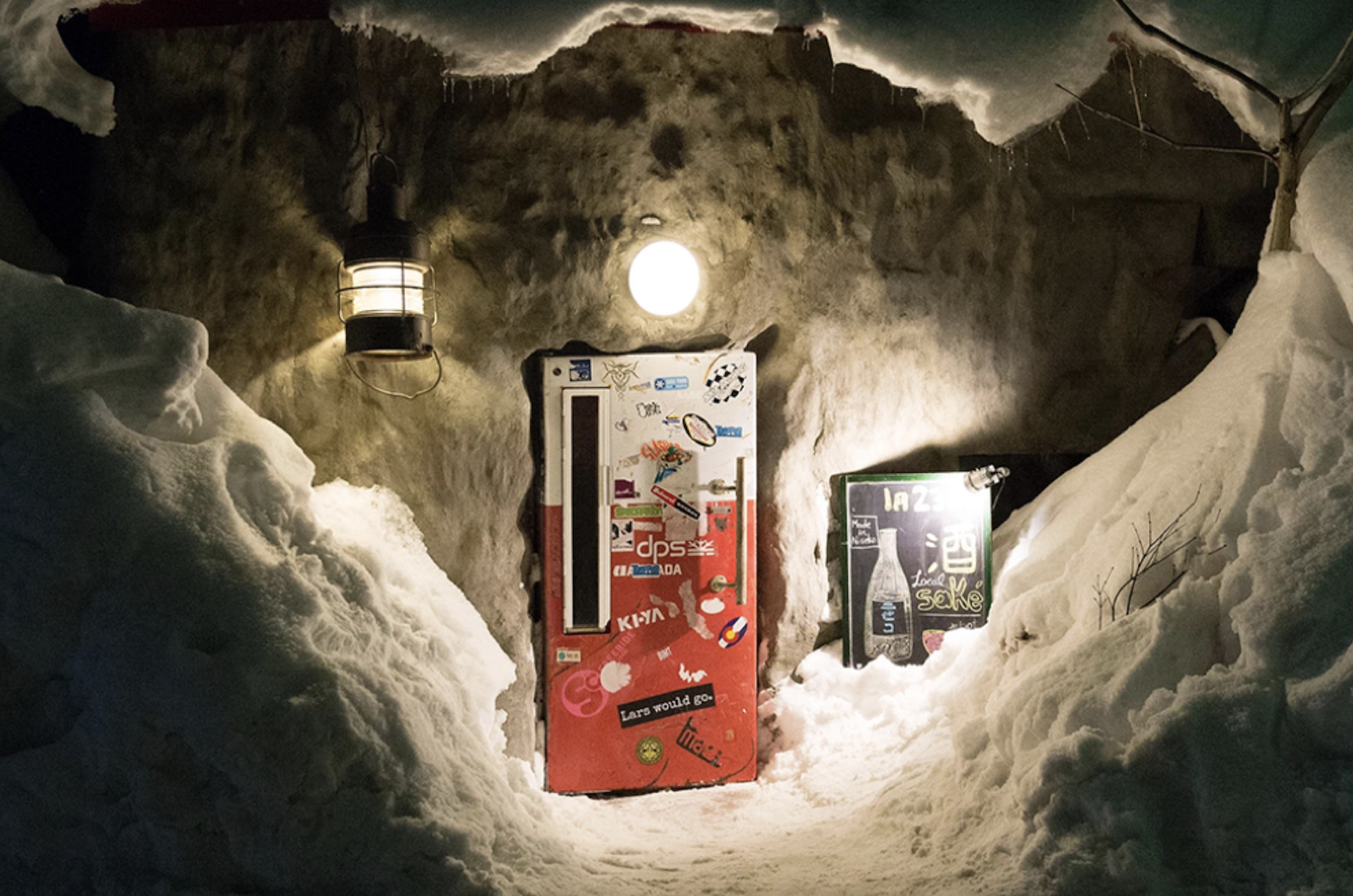 A snow-covered entrance to a building with a red door covered in stickers, lit by two round lights and surrounded by deep snow at night.