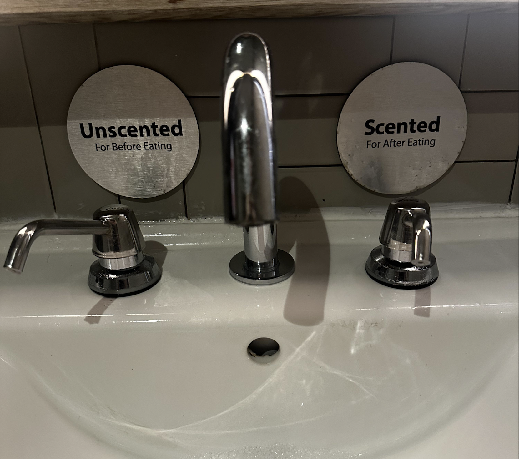 A sink with two soap dispensers labeled “Unscented for before eating” on the left and “Scented for after eating” on the right, above separate faucet handles.