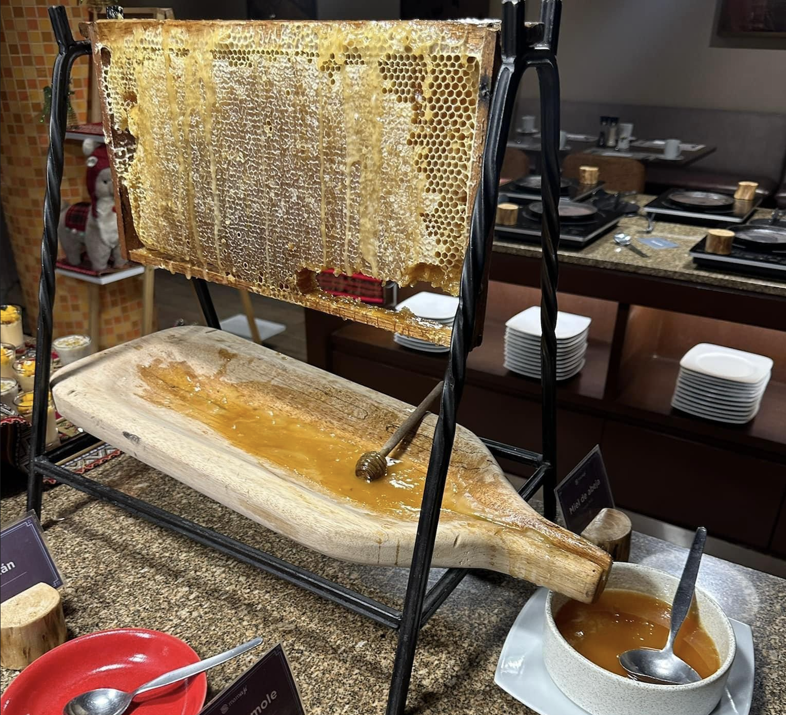 A honeycomb frame on a stand drips fresh honey onto a wooden tray with a honey dipper, next to a white bowl filled with honey, set on a buffet table with dishes in the background.