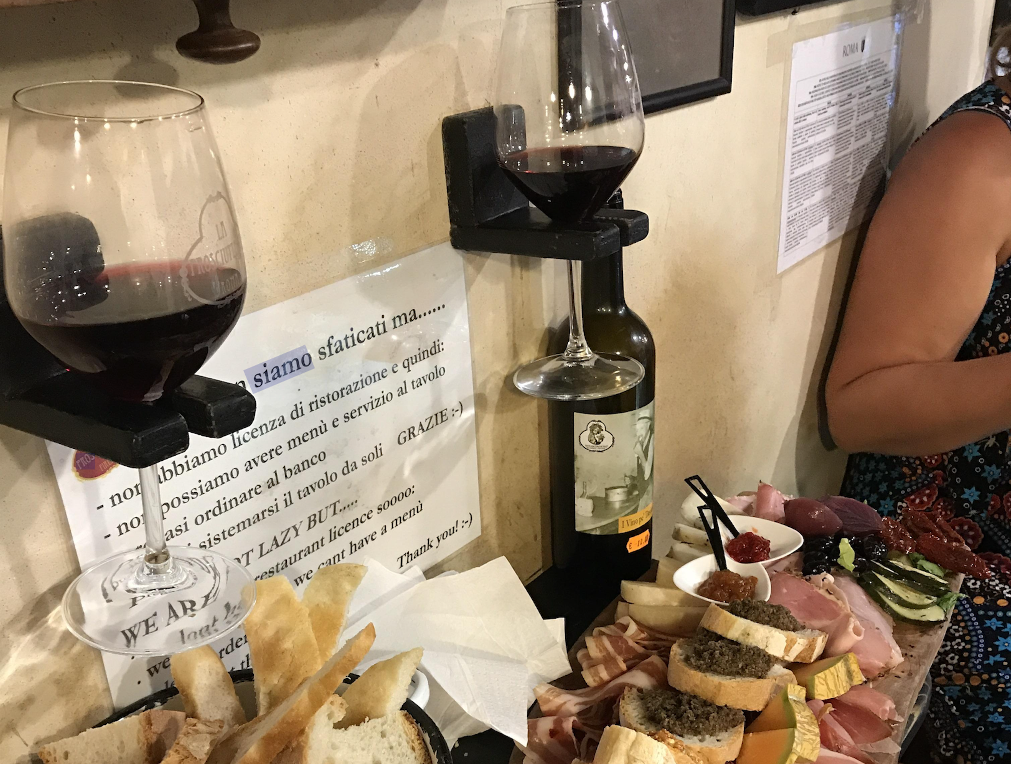 Two glasses of red wine, a bottle, assorted meats, cheeses, bread, and appetizers are arranged on a counter. A person in a floral shirt sits at the right edge. A sign with Italian and English text is visible on the wall.