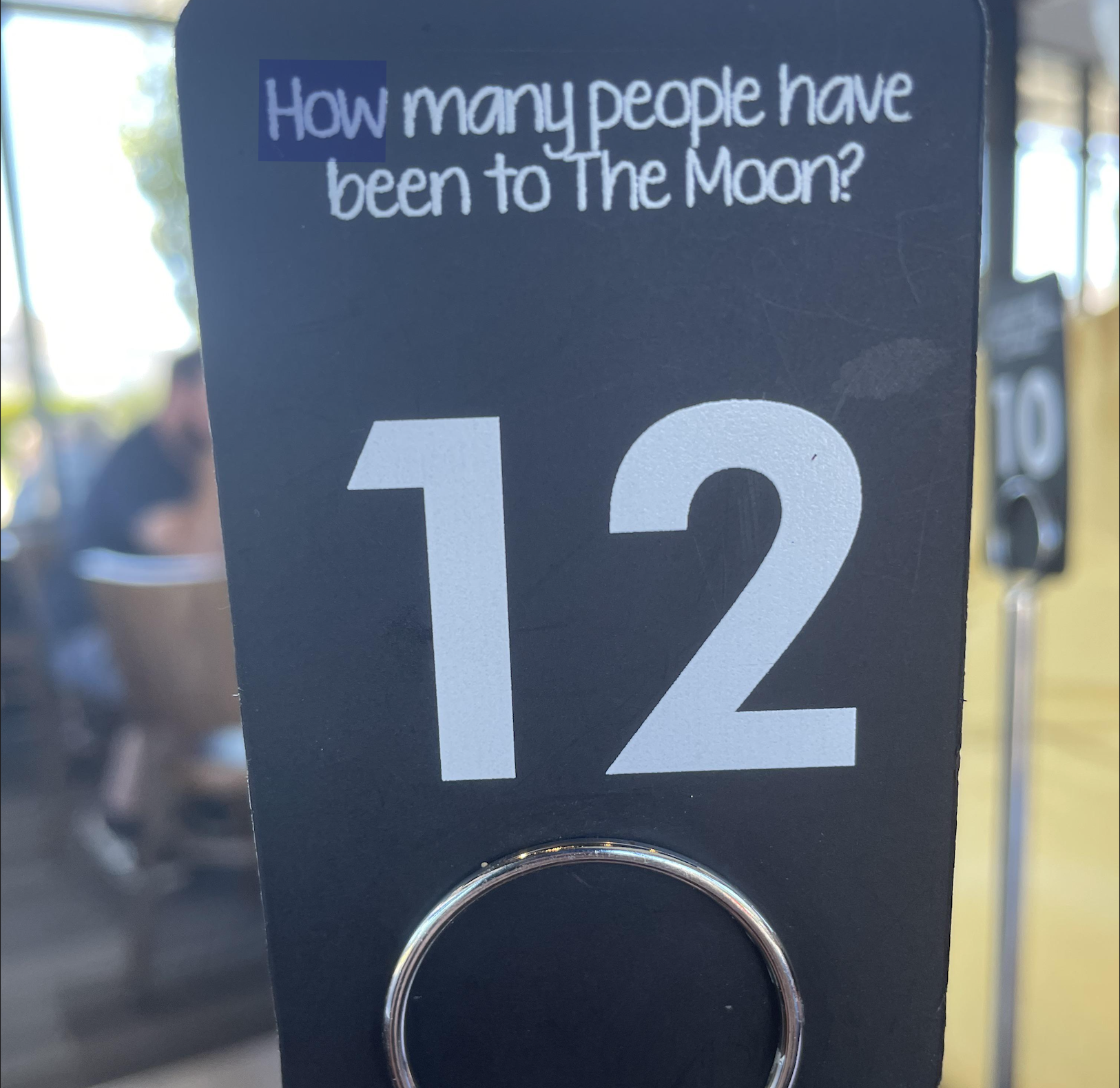 A black sign with large white number 12. At the top, it reads “How many people have been to The Moon?” in white text. A metal ring is attached at the bottom. Blurred background shows people sitting indoors.