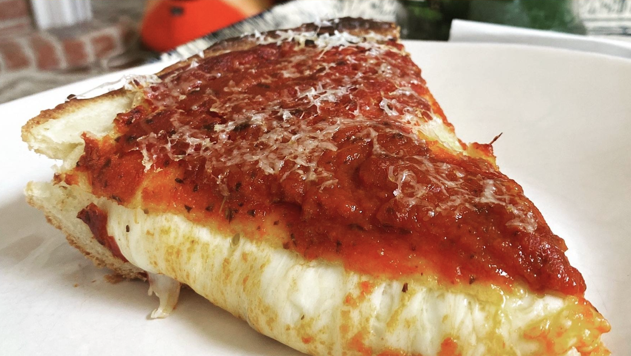 A close-up of a slice of thick-crust pizza topped with melted cheese, tomato sauce, and grated cheese on a white plate.