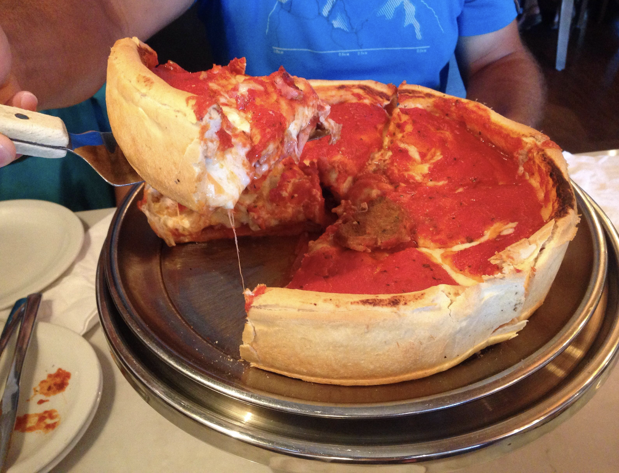 A person serves a slice of deep-dish pizza with melted cheese from a metal tray. The thick pizza has a golden crust and red tomato sauce on top.