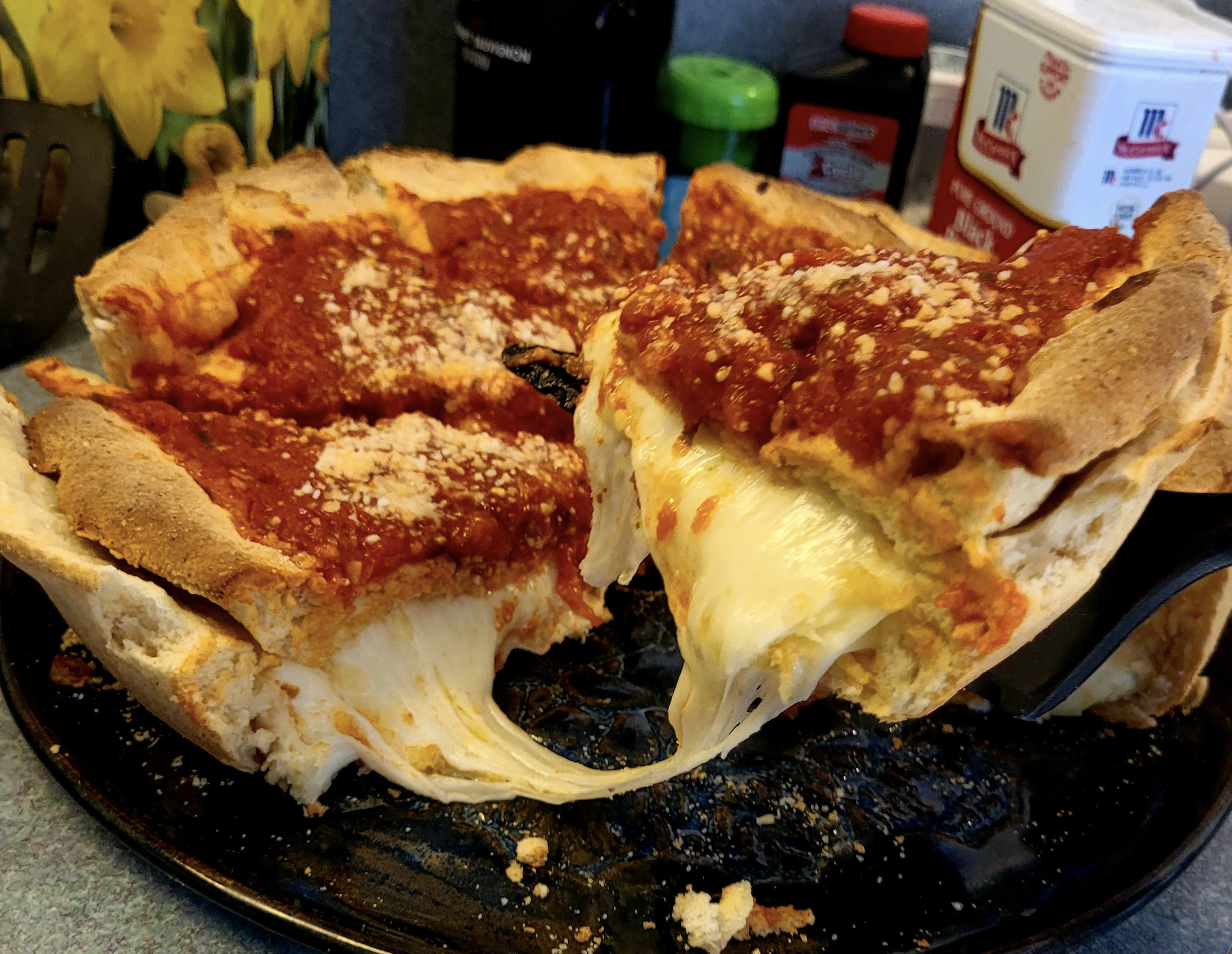 A deep-dish pizza with a thick crust, rich tomato sauce, and melted cheese is being served, showing gooey cheese stretching from the slice to the pan.
