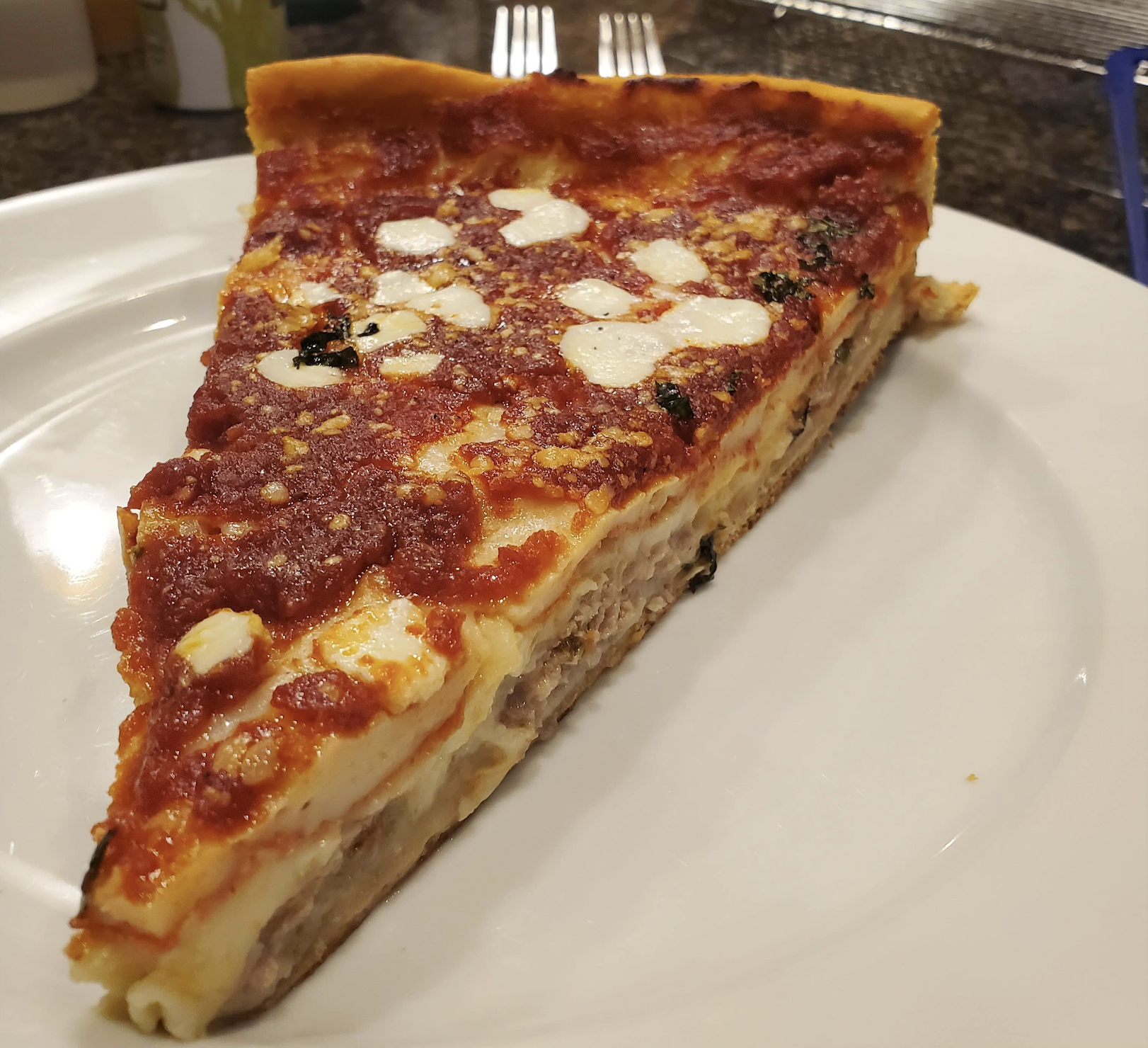 A slice of Chicago-style deep-dish pizza with tomato sauce, cheese, and sausage sits on a white plate. Two forks are visible in the background.
