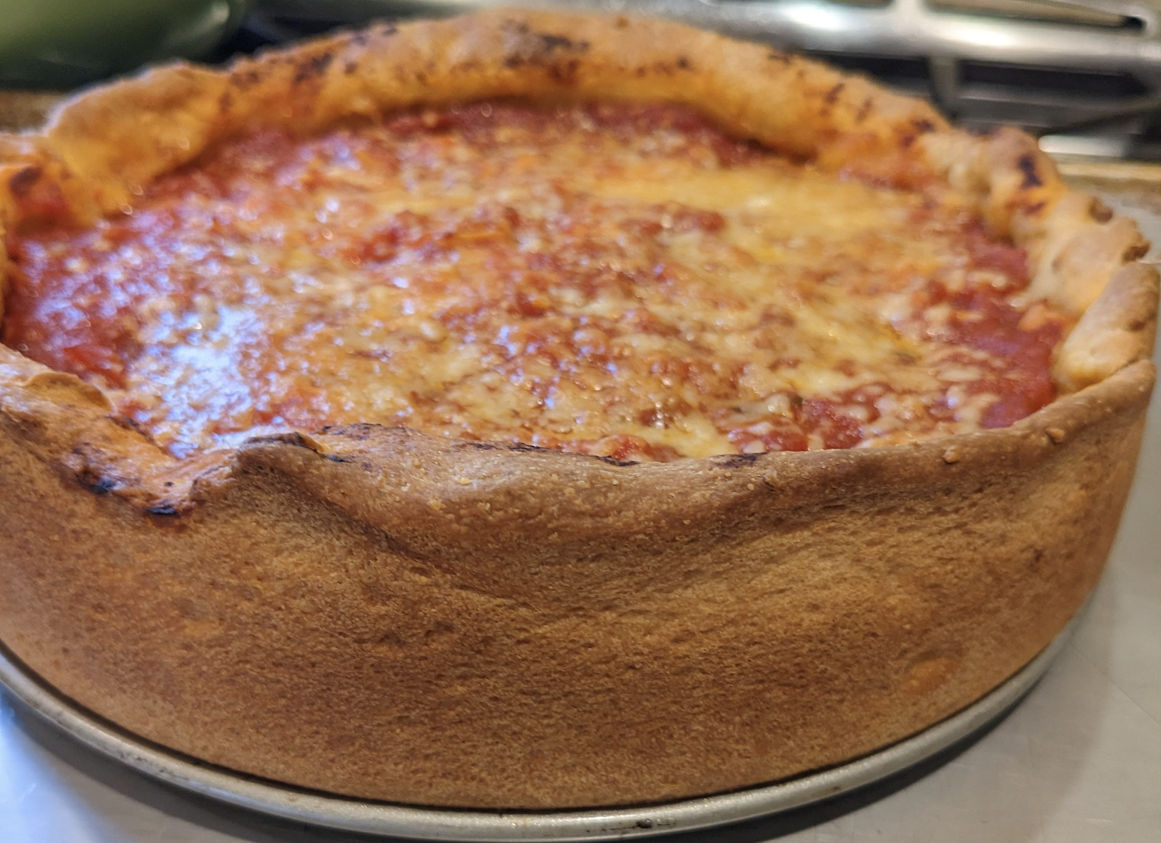 A close-up of a deep-dish pizza with a thick, golden-brown crust and a layer of melted cheese and tomato sauce on top, sitting on a metal pan.
