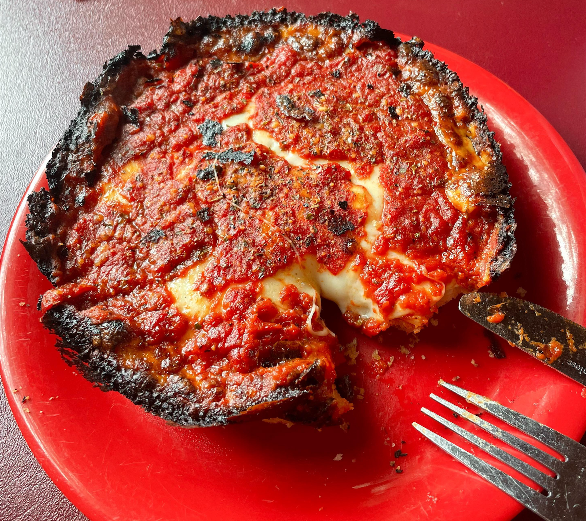 A deep-dish pizza with a heavily charred crust, melted cheese, and tomato sauce sits on a red plate. A slice has been cut, with a fork and knife resting nearby.