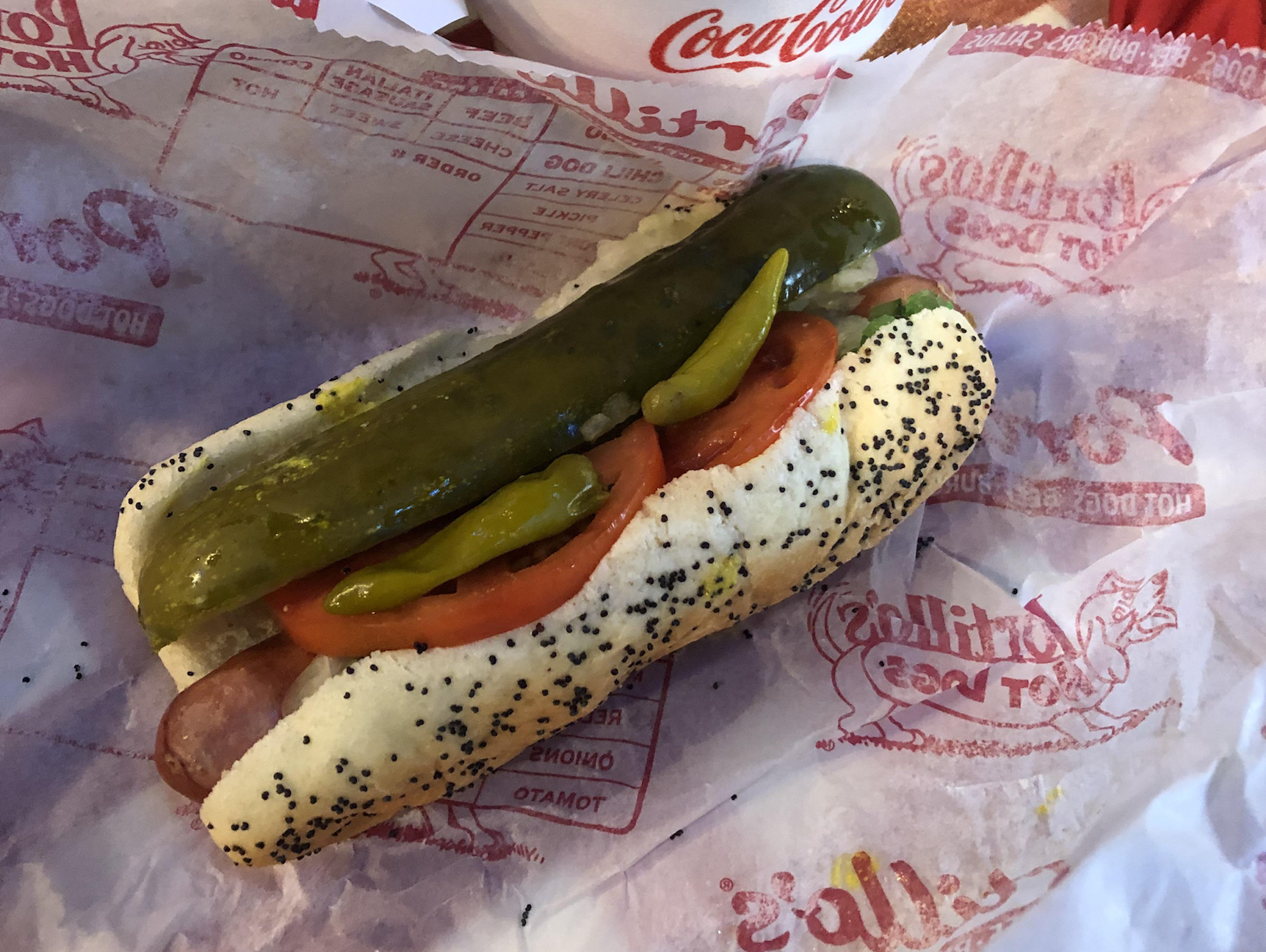 A Chicago-style hot dog in a poppy seed bun, topped with a pickle spear, tomato slices, sport peppers, and relish, on branded Portillo’s paper.