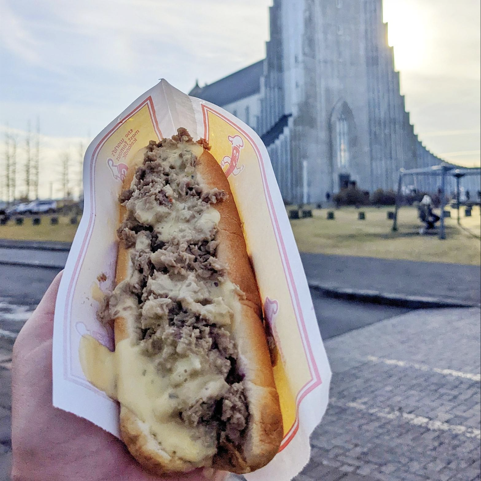 A hand holds a hot dog topped with melted cheese and meat, in front of the Hallgrímskirkja church in Reykjavik, Iceland, on a bright day.
