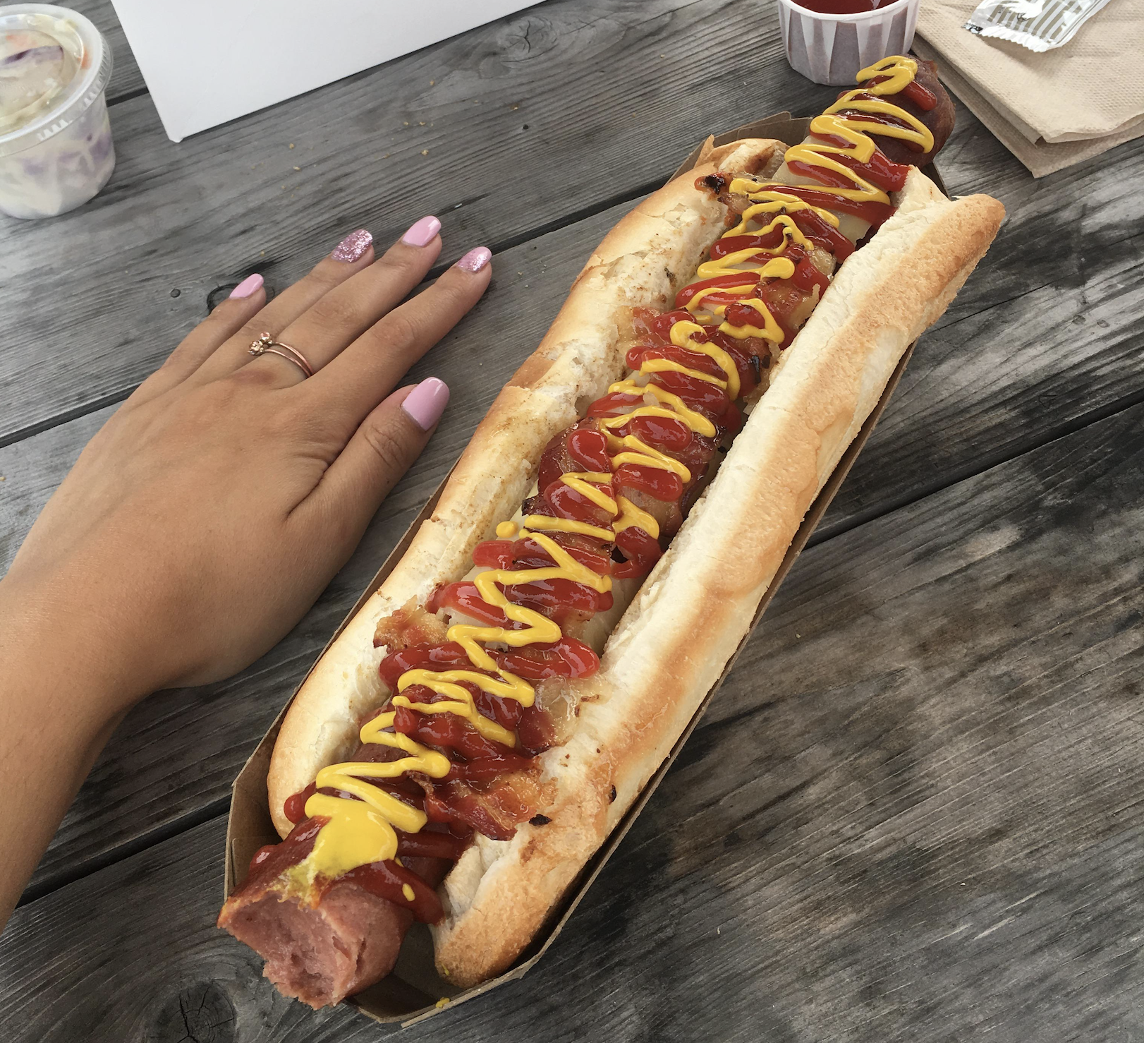 A very large hot dog topped with ketchup and mustard sits in a bun on a wooden table. A hand with pink nails rests next to it for size comparison.