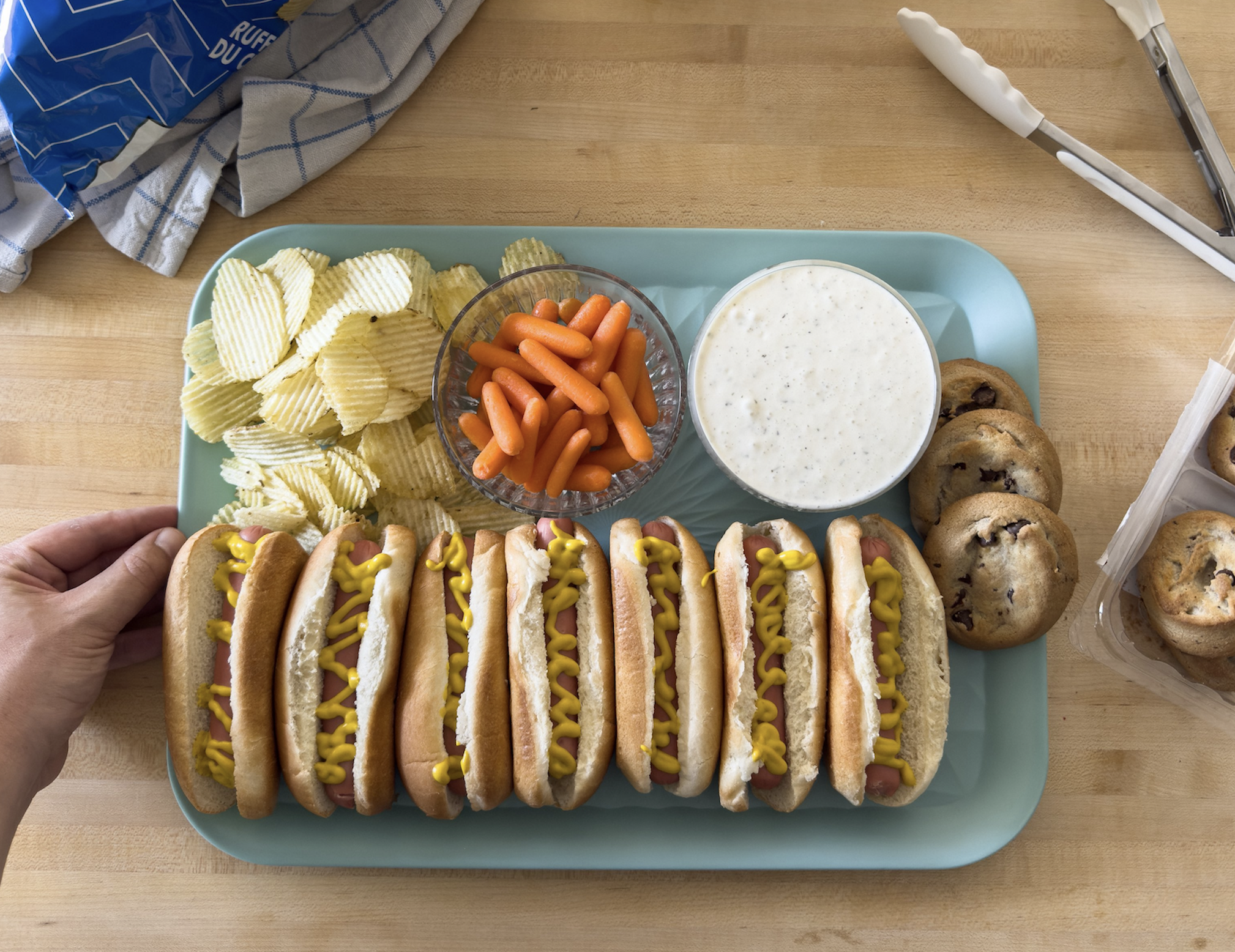 A tray with seven hot dogs topped with mustard, potato chips, baby carrots with ranch dip, and chocolate chip cookies that demonstrate hot dogs variations. A hand holds the tray on a wooden table with tongs and chips in the background.