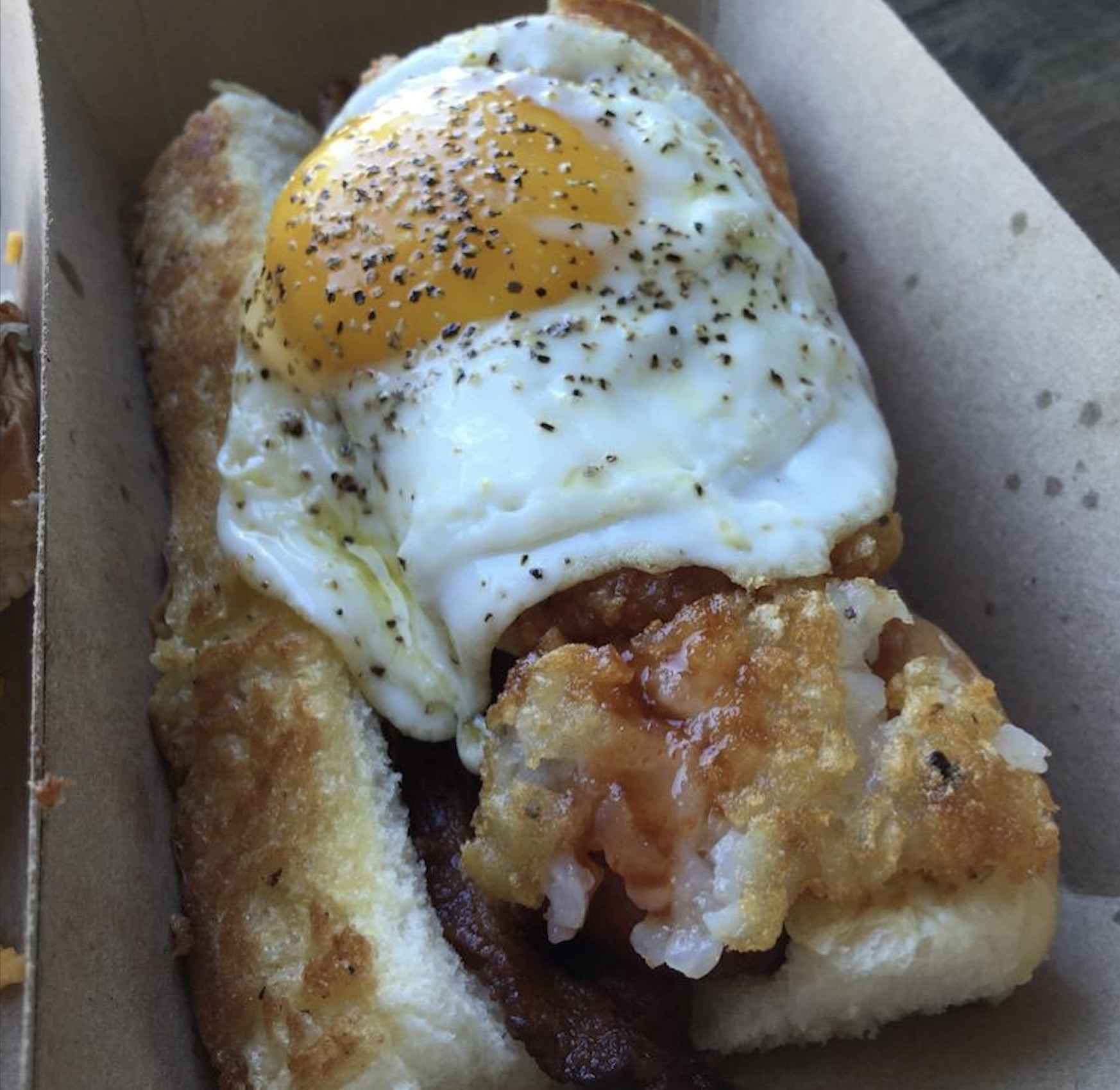 A sandwich on a soft bun topped with a sunny-side-up egg, ground black pepper, crispy hash browns, and bacon, served in a cardboard container.