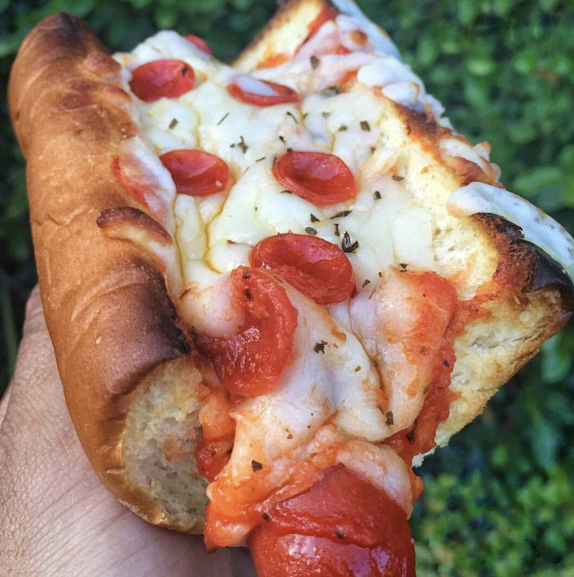A close-up of a hand holding a pizza-stuffed hot dog bun topped with melted cheese, pepperoni slices, and herbs, with a bite taken out of one end.