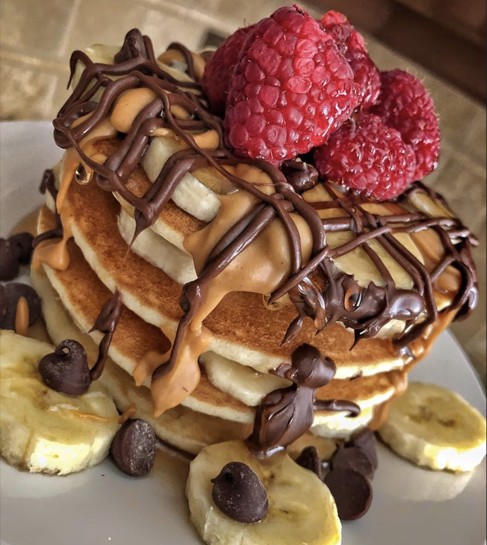 A stack of pancakes topped with banana slices, fresh raspberries, chocolate chips, and drizzles of chocolate and peanut butter.