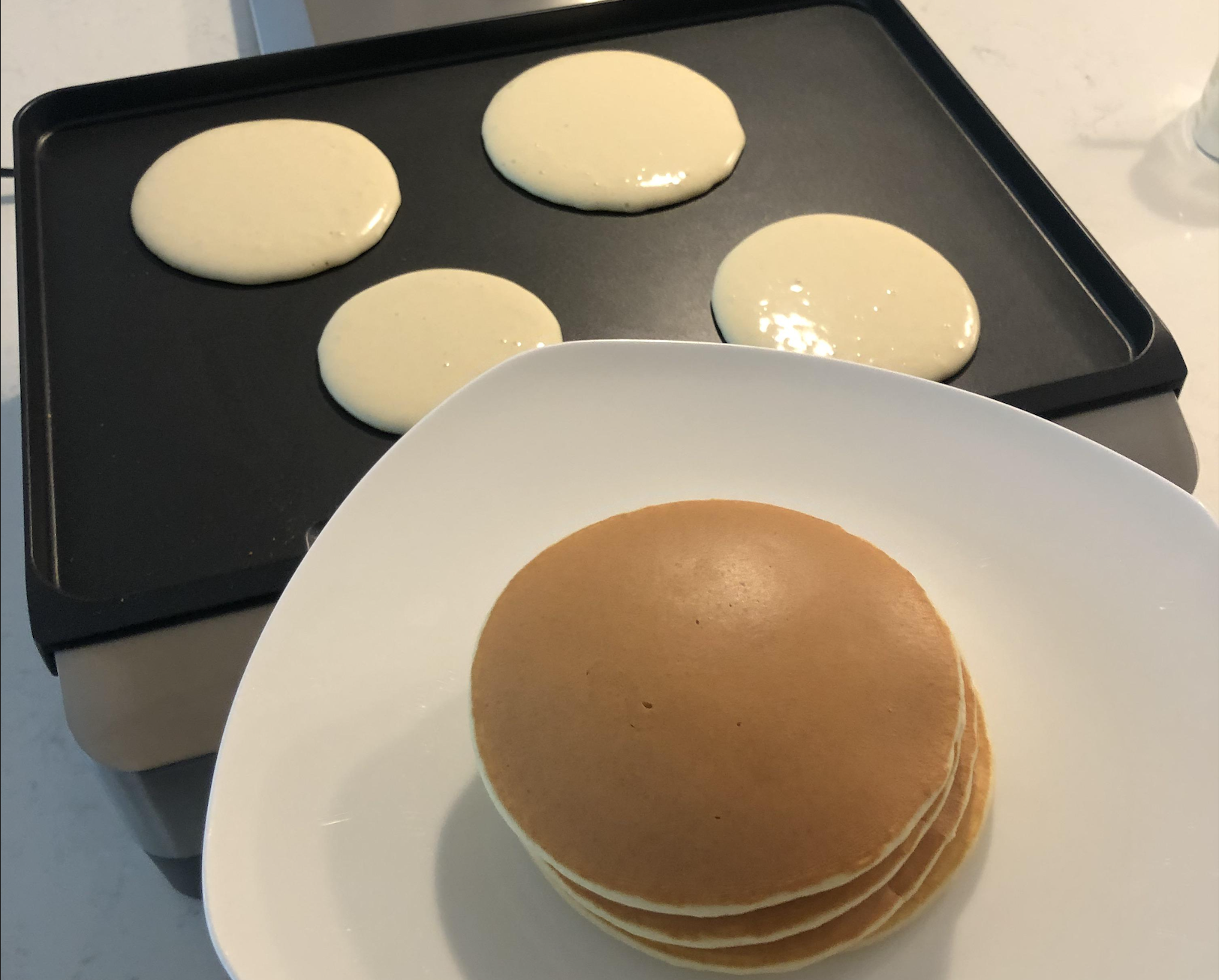 A stack of four golden-brown pancakes on a white plate in the foreground, with four more pancakes cooking on a black griddle in the background.