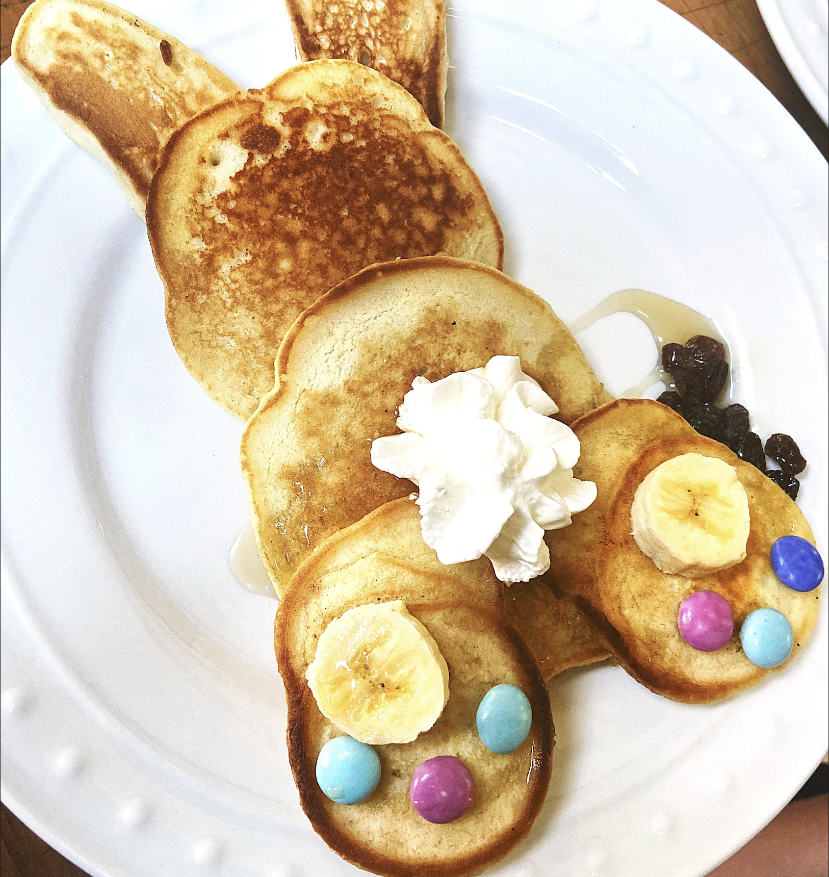 Pancakes arranged on a plate to look like a rabbit, decorated with whipped cream, banana slices, and colorful candy pieces.