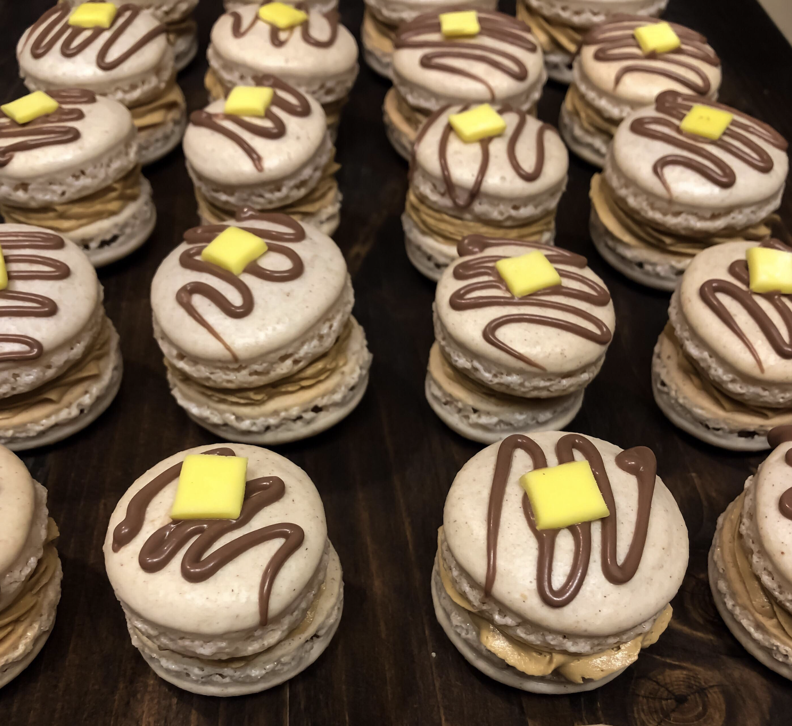 A tray of pancake-themed macarons, each topped with a chocolate drizzle and a small yellow candy square.