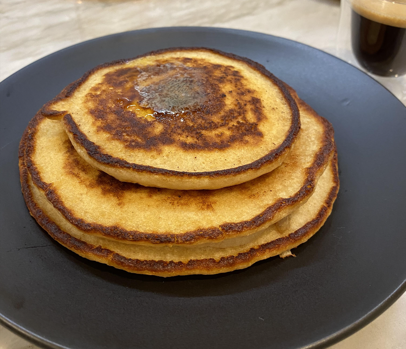 A stack of three golden-brown pancakes with a pat of melting butter on top, served on a black plate next to a glass of espresso.