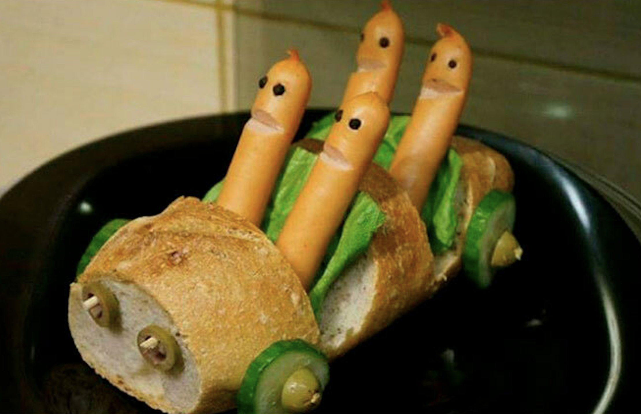 A baguette carved into the shape of a car with four hot dog figures as passengers and cucumber slice wheels.