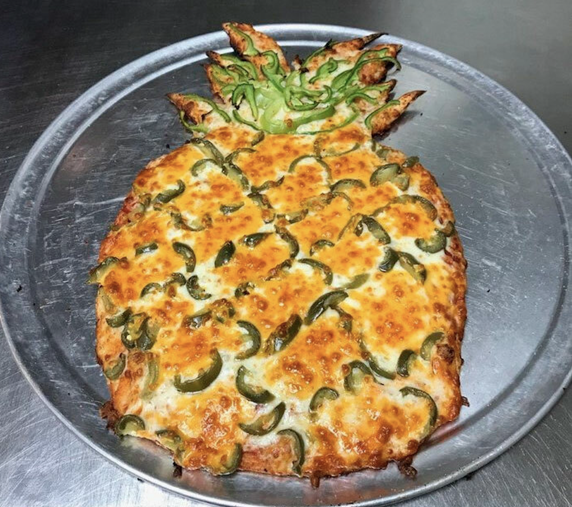 Pineapple-shaped pizza topped with cheese and sliced jalapeños on a metal tray, with green bell pepper strips forming the pineapple crown.