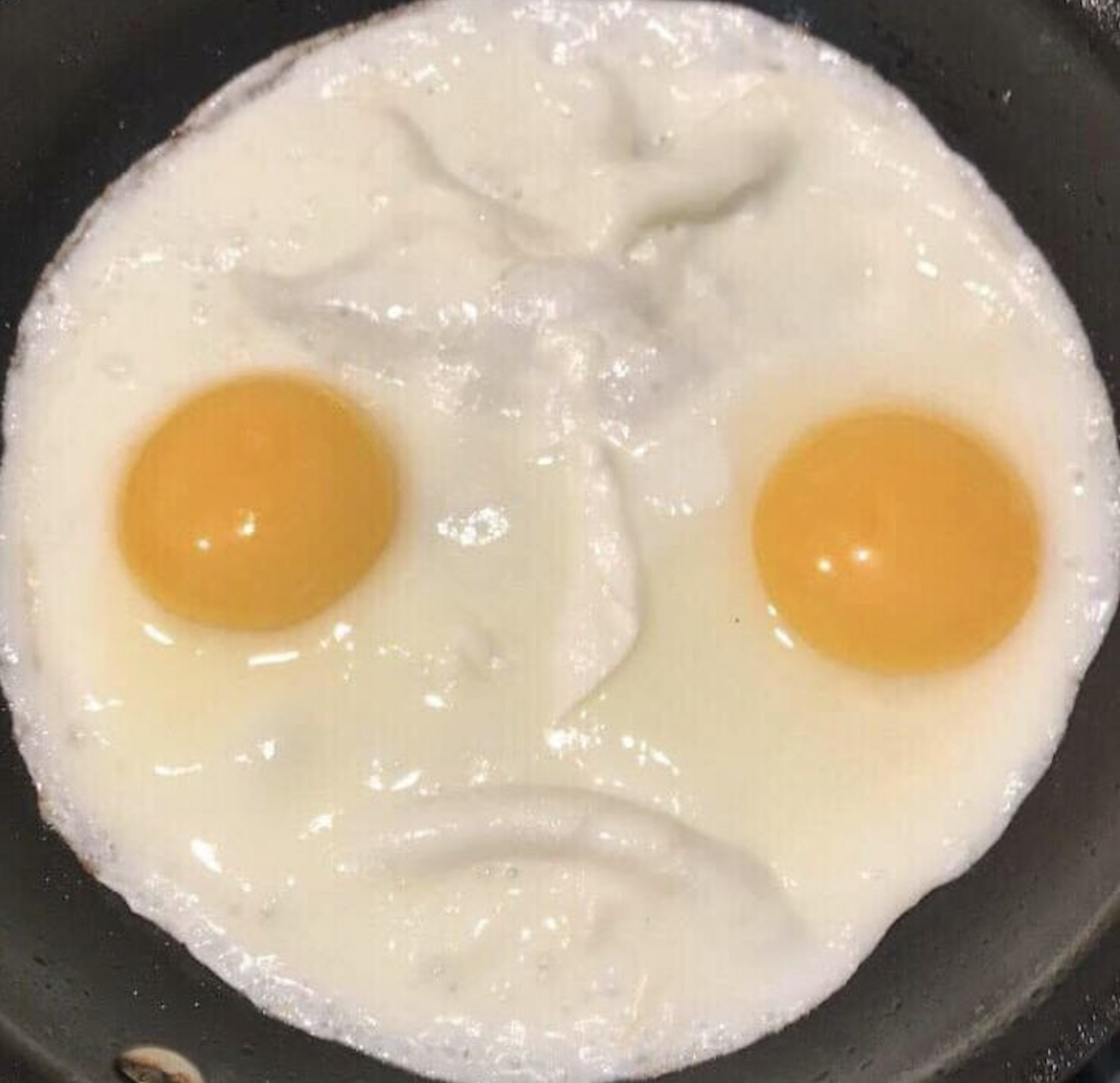 Two fried eggs in a skillet, with the yolks and egg whites arranged to look like a sad face.