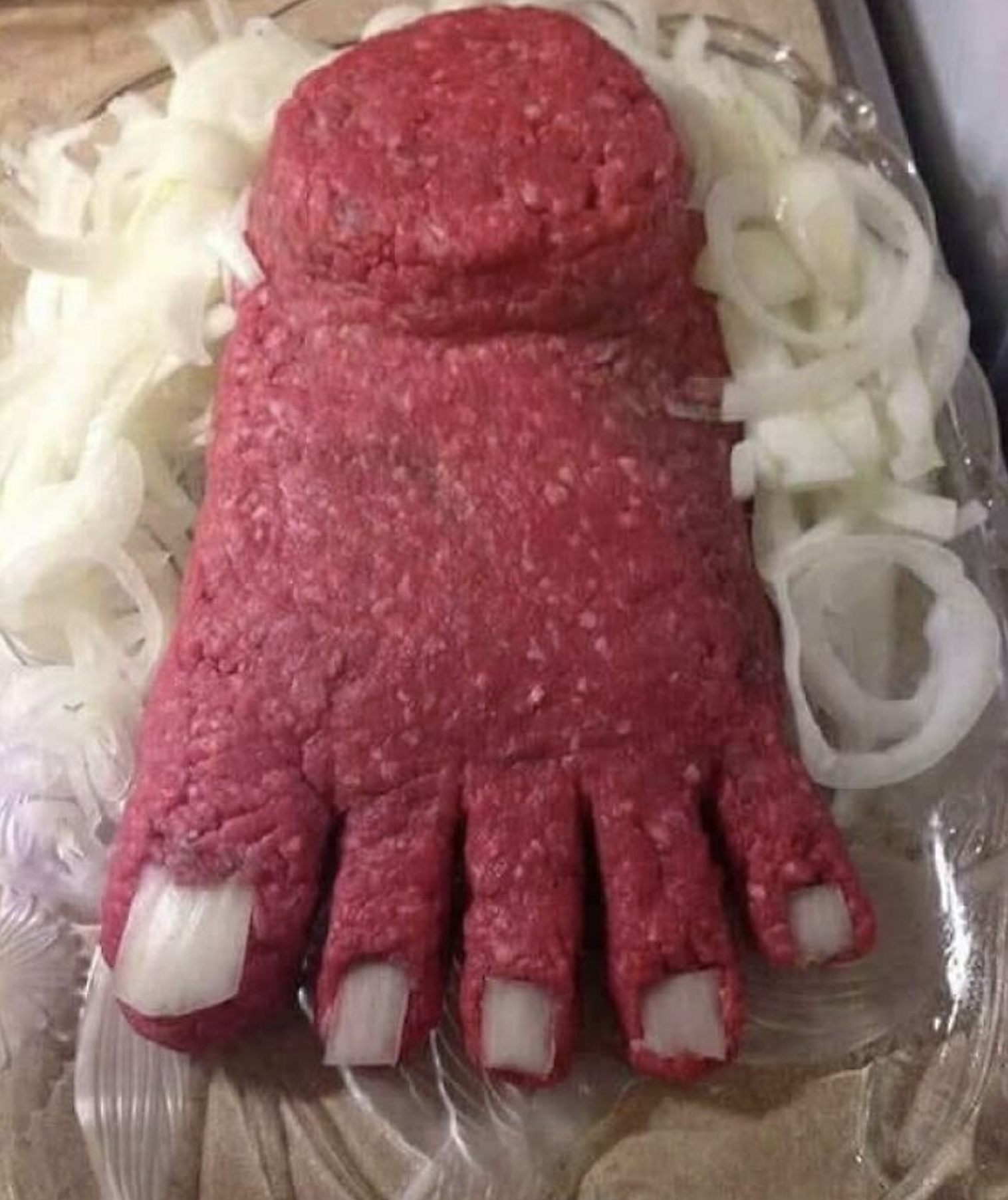 Ground beef shaped like a human foot, with onion slices serving as toenails, surrounded by sliced onions on a plate.