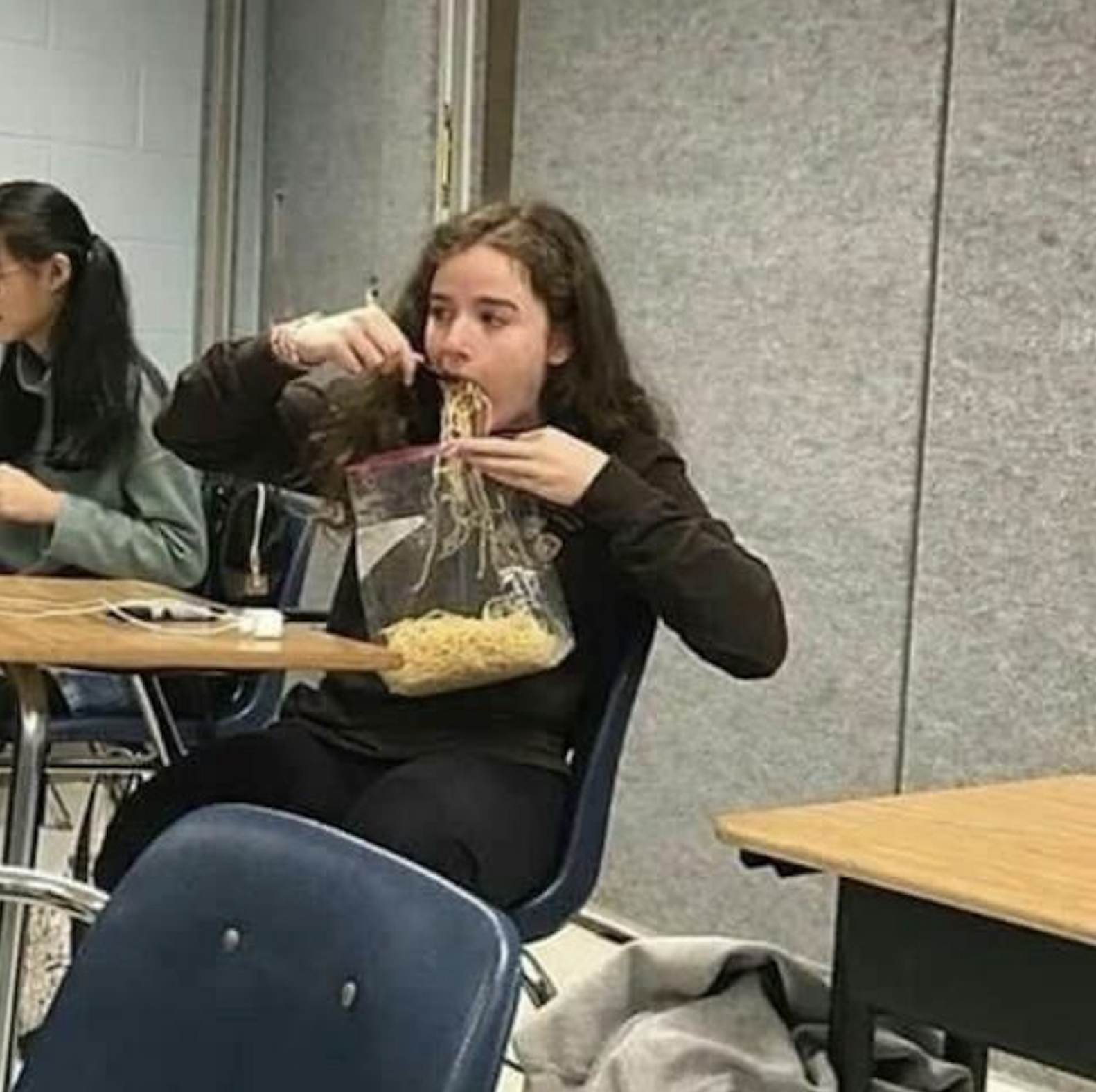 A student sitting at a desk eating spaghetti directly from a plastic storage bag.