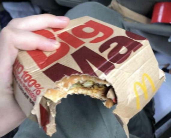 A person holds a McDonald’s burger box that has been bitten into, through both the cardboard packaging and the sandwich inside.